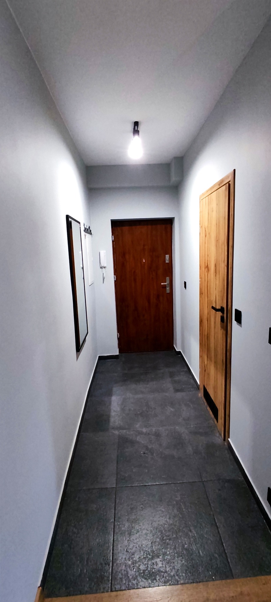 Wnętrze korytarza po odświeżeniu: szare ściany, ciemna, kwadratowa podłoga z płytek, lustro w czarnej ramie, drewniane drzwi po prawej stronie i ciemnobrązowe drzwi wejściowe na wprost.
