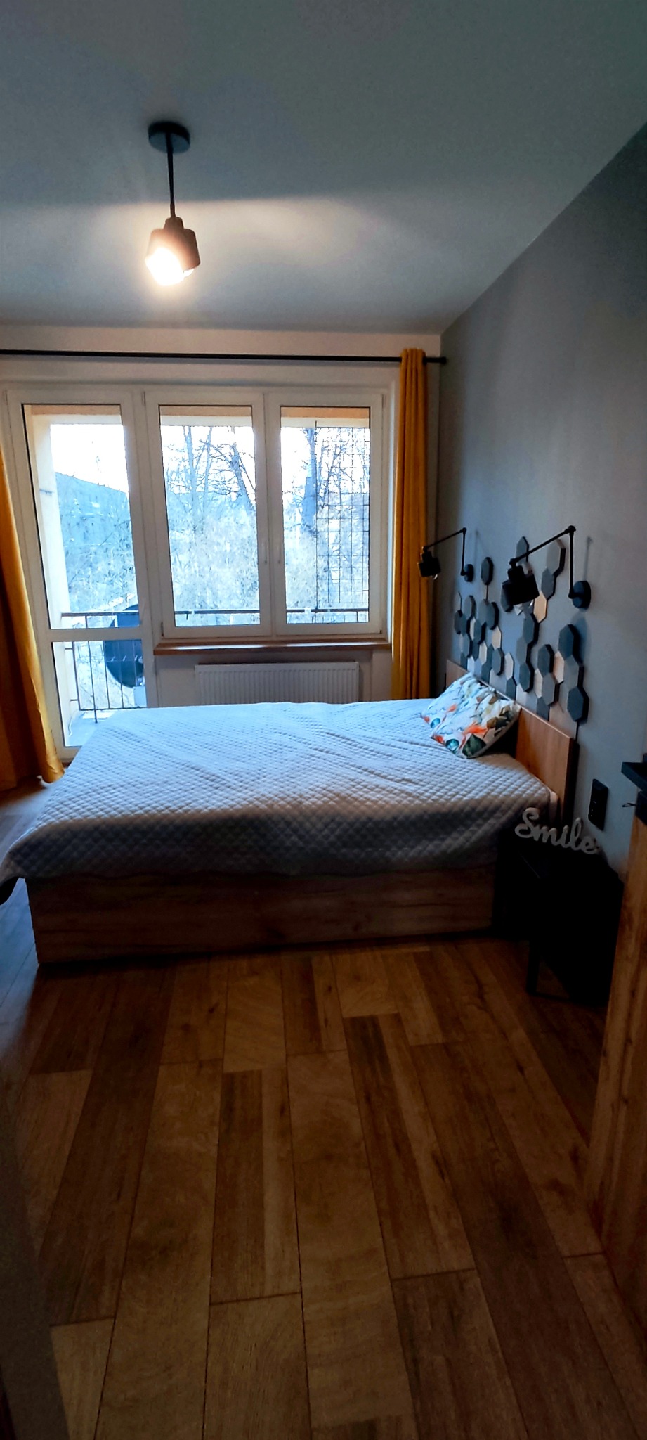 Sypialnia w szaro-żółtej tonacji z drewnianą podłogą i łóżkiem z dekoracyjnym zagłówkiem z heksagonów, oświetlona lampami ściennymi i sufitową, z widokiem na balkon przez okno.