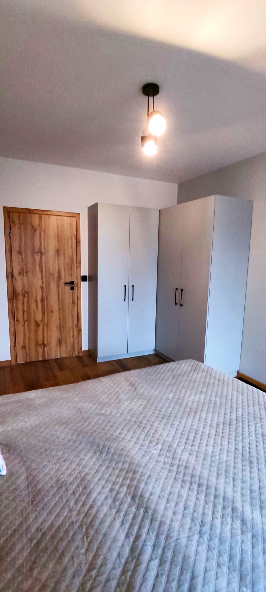 Jasne wnętrze sypialni z drewnianą podłogą, łóżkiem z pikowaną narzutą, białymi szafami w zabudowie i drewnianymi drzwiami z czarną klamką, oświetlone nowoczesną lampą sufitową.