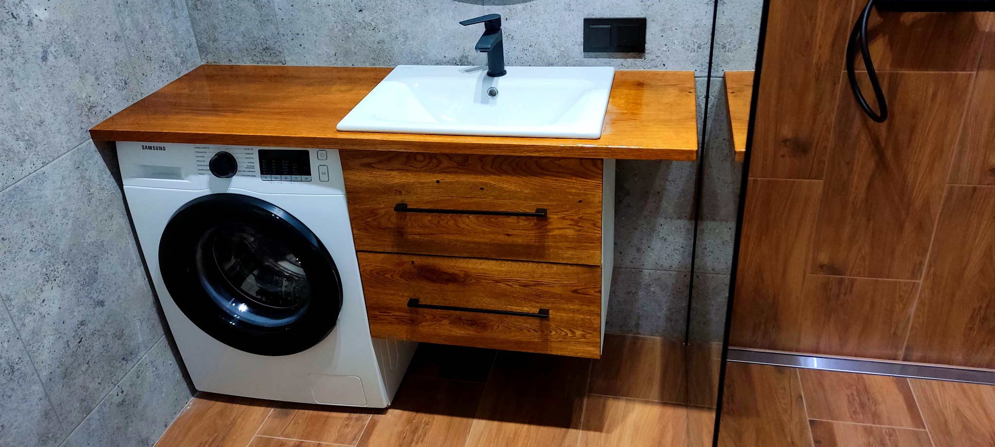 Nowoczesna łazienka z drewnianym blatem i szafką pod umywalkę, obok pralka Samsung, szare płytki na ścianie i drewnopodobne na podłodze, czarna bateria umywalkowa i włącznik światła.