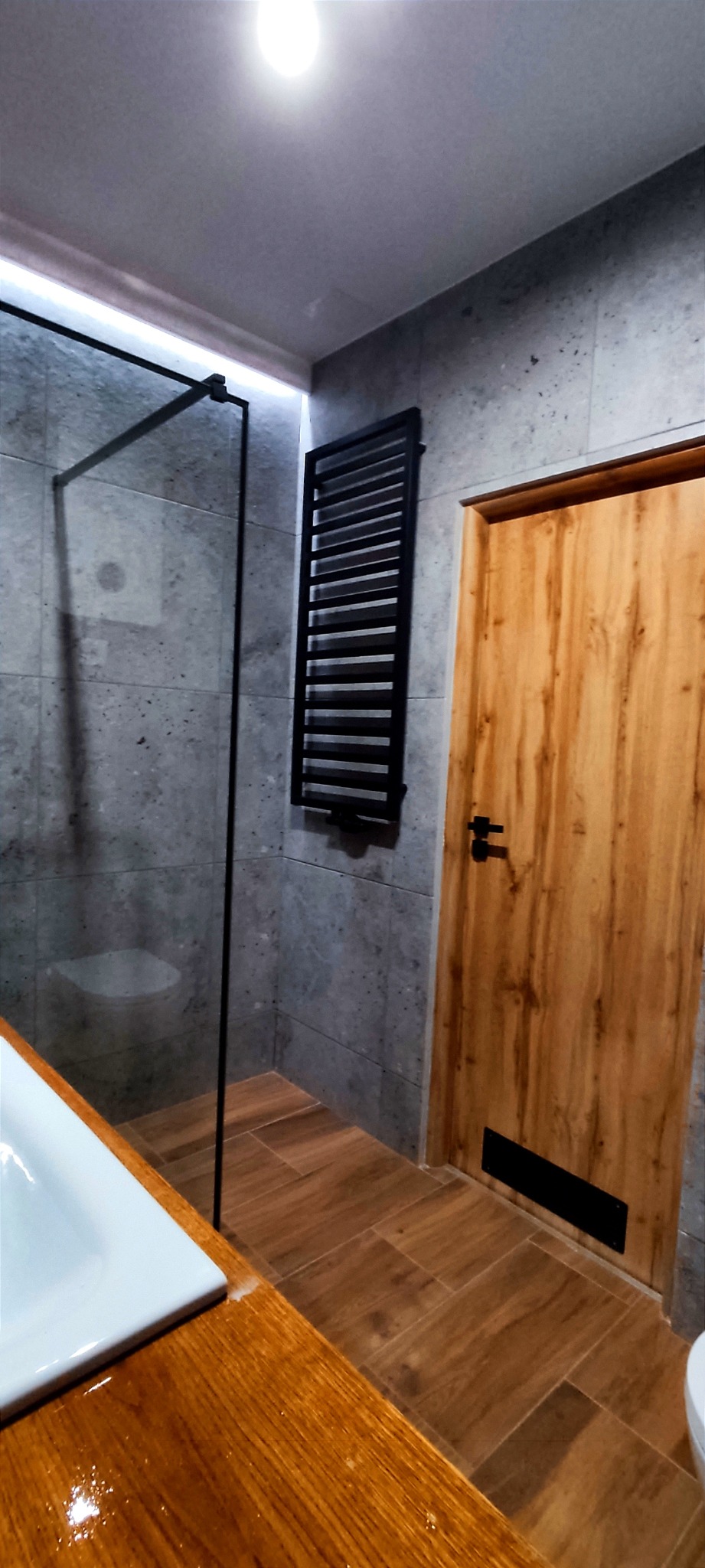Nowoczesna łazienka z szarymi płytkami imitującymi kamień, drewnopodobną podłogą i akcentami w czerni: grzejnik drabinkowy, rama kabiny prysznicowej, detale drzwi.