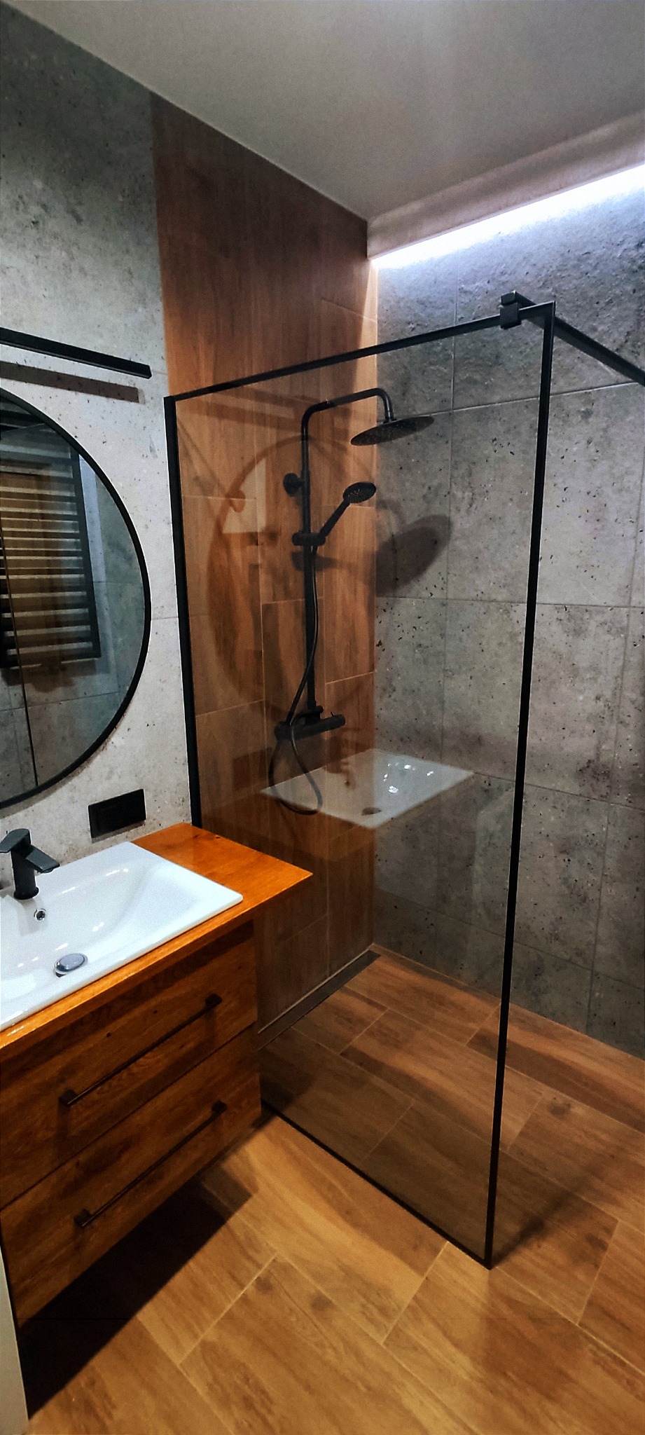 Nowoczesna łazienka z kabiną prysznicową typu walk-in z czarnymi okuciami, drewnianą szafką pod umywalką z blatem z litego drewna i okrągłym lustrem.