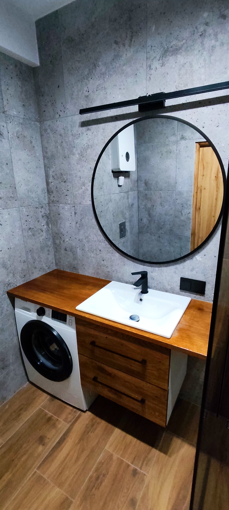 Nowoczesna łazienka z pralką zabudowaną pod drewnianym blatem umywalki, okrągłe lustro z czarną ramą, szare płytki na ścianach i brązowe płytki imitujące drewno na podłodze.