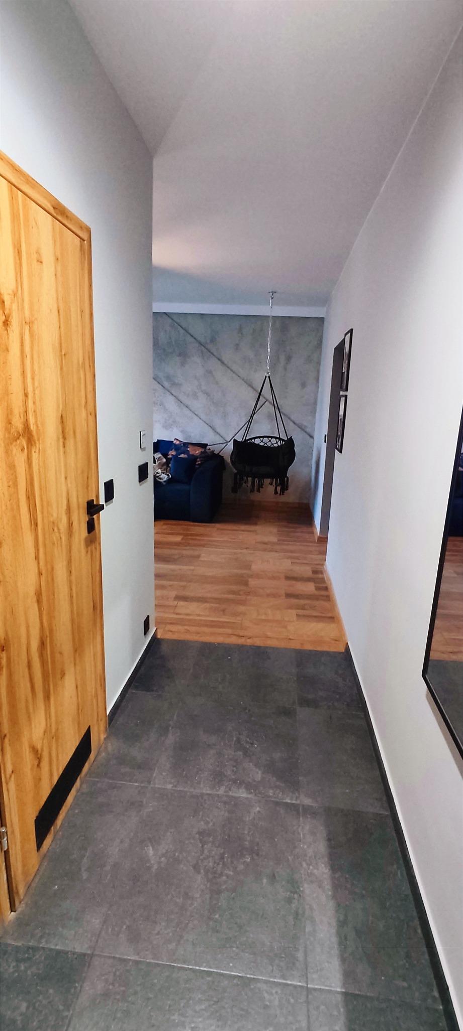 Wnętrze mieszkania z ciemnoszarą podłogą w korytarzu przechodzącą w drewnianą podłogę w salonie, widoczny hamak wiszący pod sufitem i ściana z betonową fakturą.