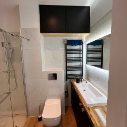 Nowoczesna łazienka z kabiną prysznicową, podwieszaną toaletą i umywalką nablatową na drewnianym blacie, oświetlona lustrem z ledowym podświetleniem i punktowym światłem sufitowym.