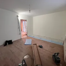 Pokój w trakcie układania paneli podłogowych, widoczne panele w opakowaniu, podkład, narzędzia i odkurzacz przemysłowy, ściany przygotowane do malowania.