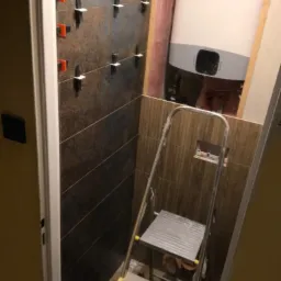 Proces układania ciemnych, podłużnych płytek w łazience, widoczne klipsy poziomujące, drabina aluminiowa i bojler elektryczny na ścianie.