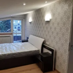 Pokój z łóżkiem w folii ochronnej, ściana w jasne, wzorzyste tapety, drewniana podłoga, lampki ścienne i okno z widokiem na zimowy krajobraz.