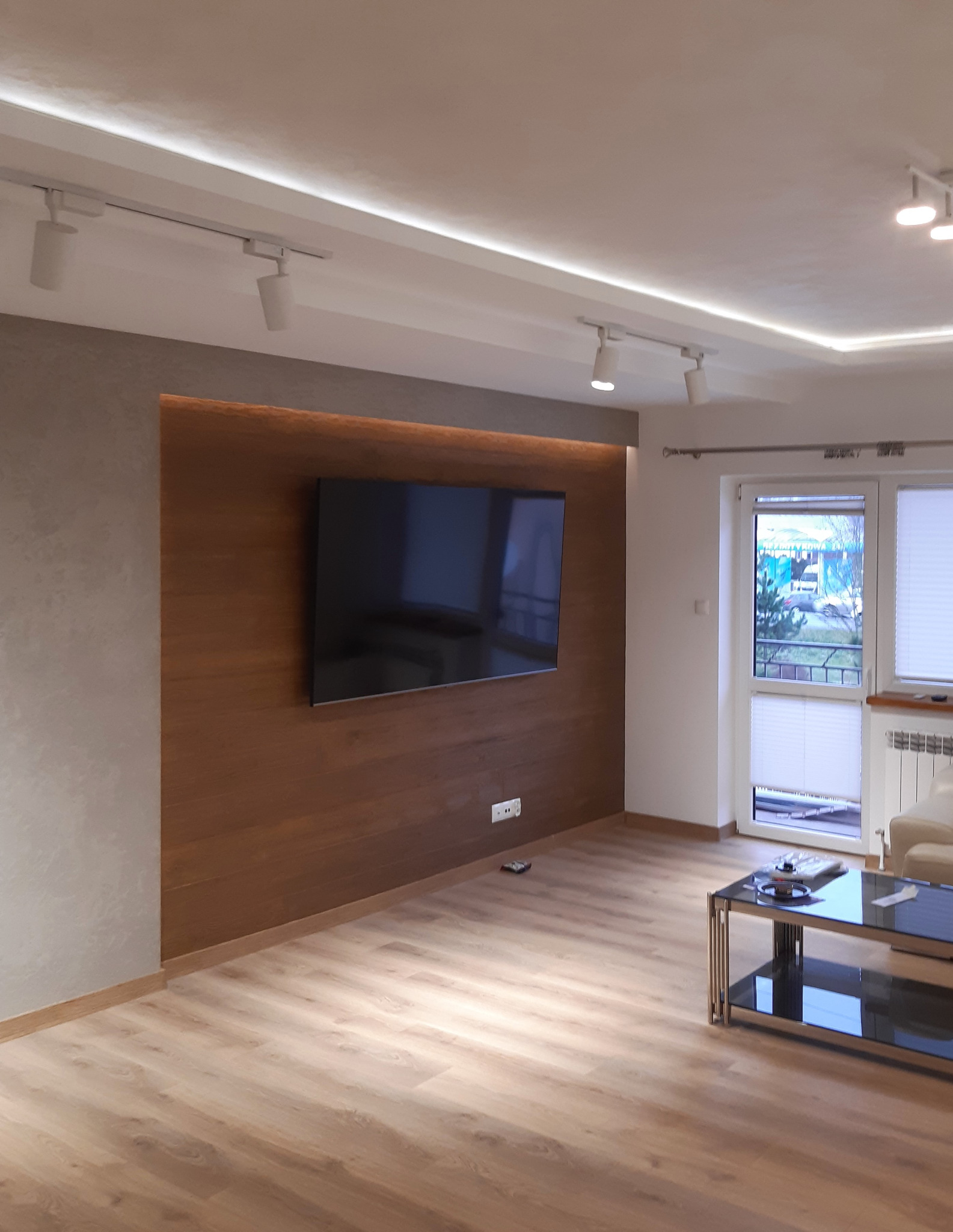 Nowoczesny salon z drewnianą ścianą akcentującą, na której wisi telewizor, parkiet w odcieniach beżu, oświetlenie punktowe i listwa LED w suficie podwieszanym.