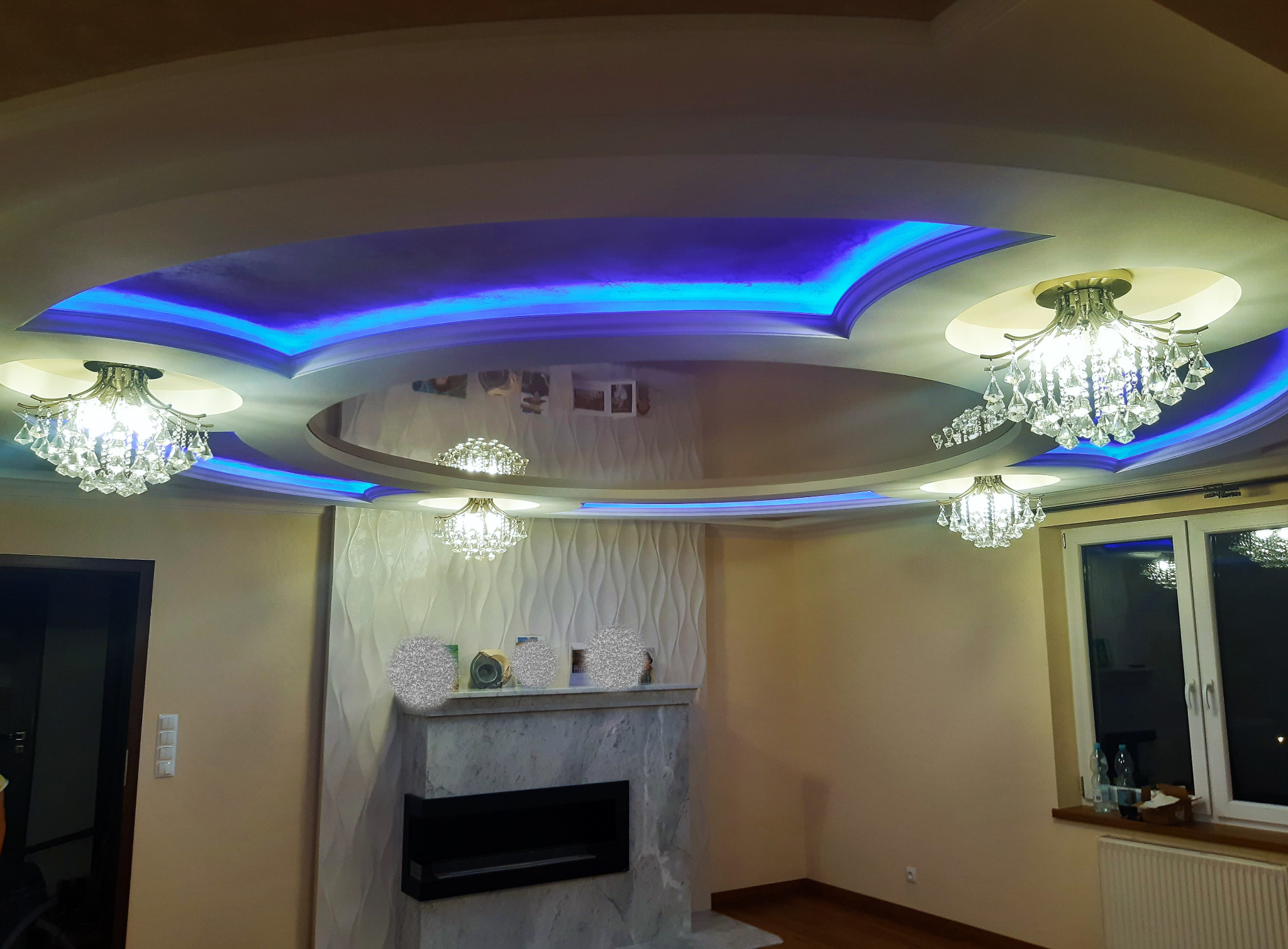 Salon z podwieszanym sufitem z niebieskim oświetleniem LED, lustrzanym panelem centralnym i kryształowymi żyrandolami, dekoracyjny kominek z fakturą 3D na ścianie.