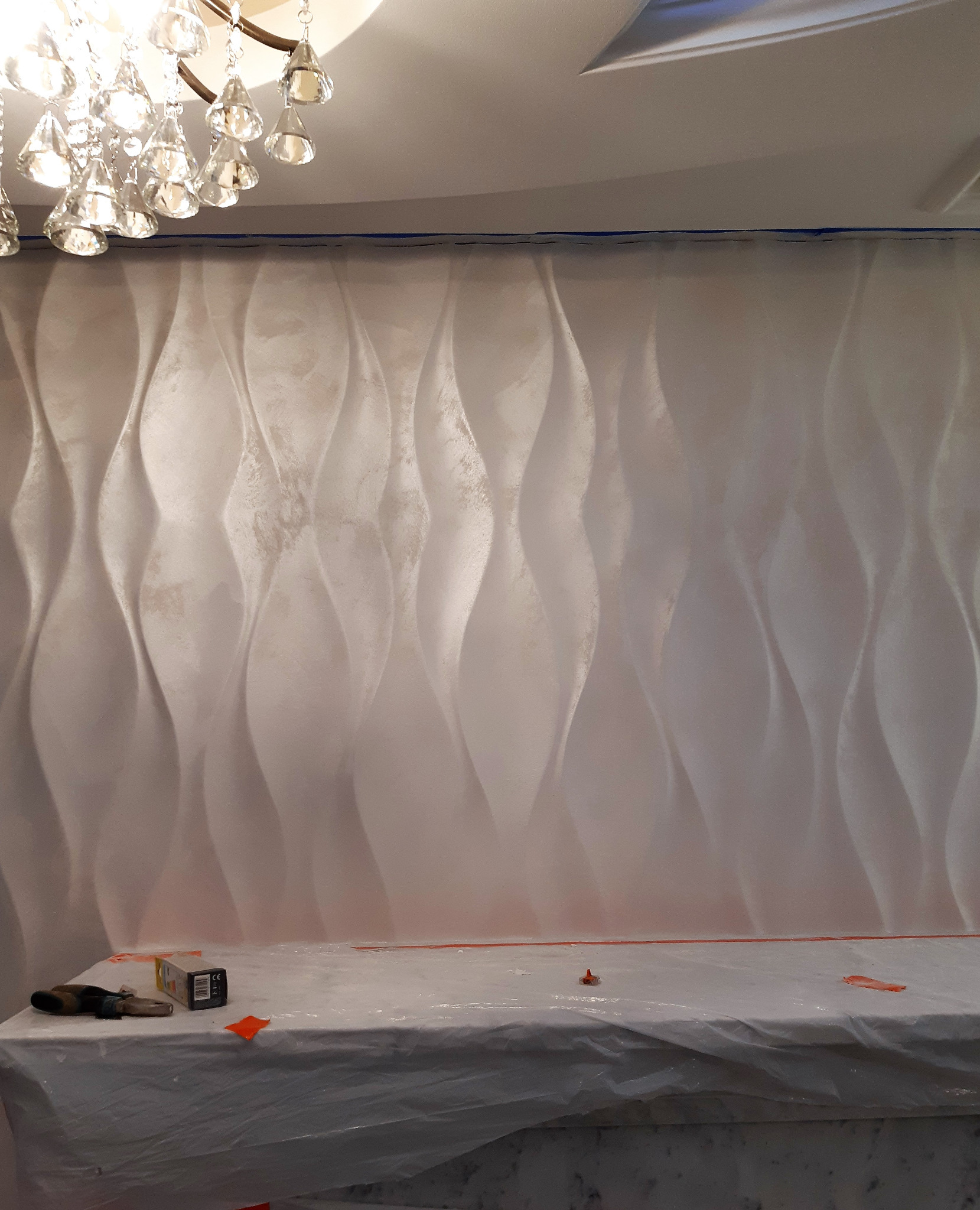 Ściana z dekoracyjnymi panelami 3D o falistym wzorze, zabezpieczona niebieską taśmą malarską u góry, z widocznym oświetleniem sufitowym i zakrytą powierzchnią roboczą poniżej, na której leżą...