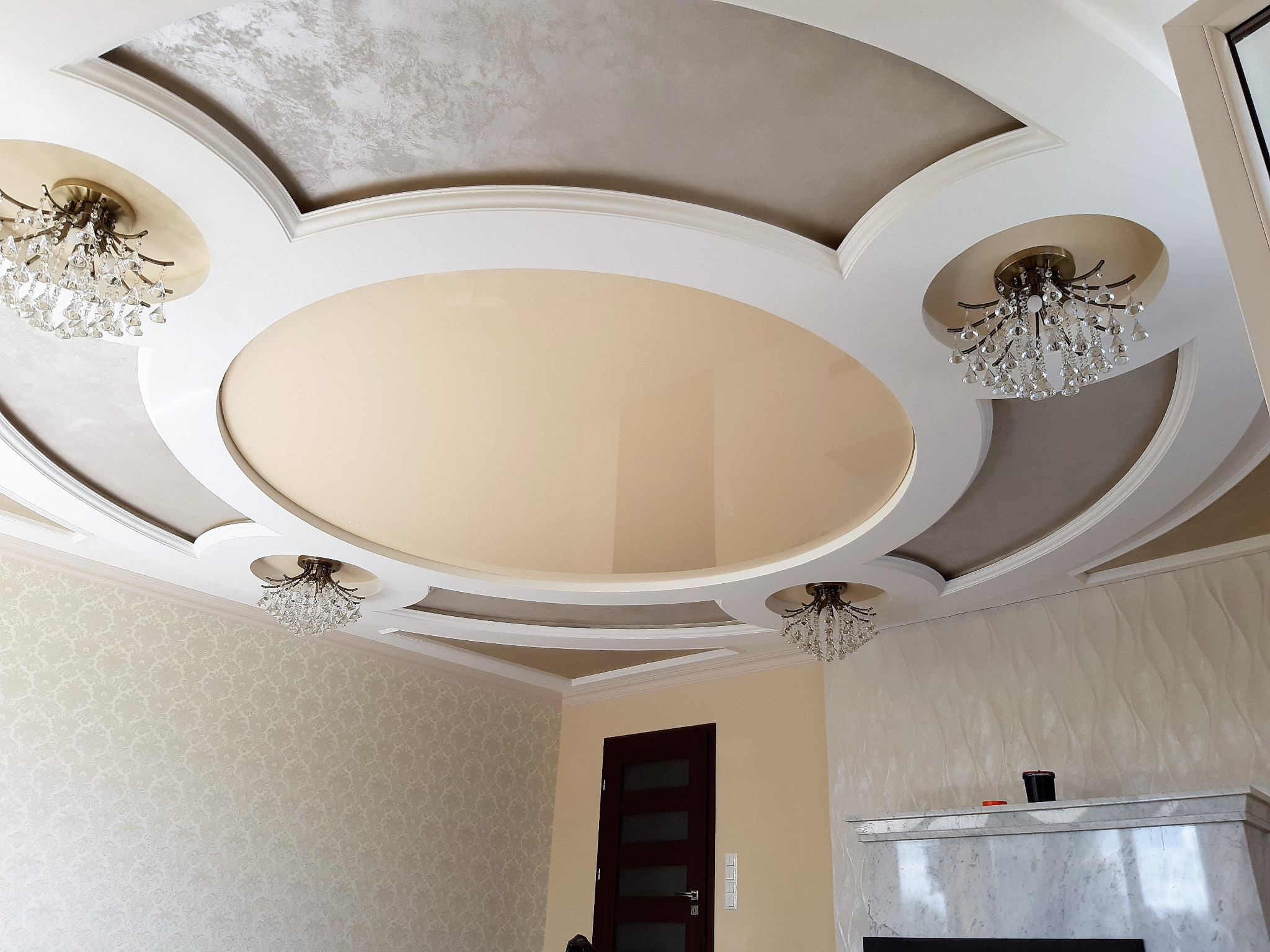 Dekoracyjny sufit wielopoziomowy z okrągłym centralnym elementem w odcieniach beżu i szarości, oświetlony czterema kryształowymi żyrandolami, z widoczną tapetą w subtelny wzór na ścianie...