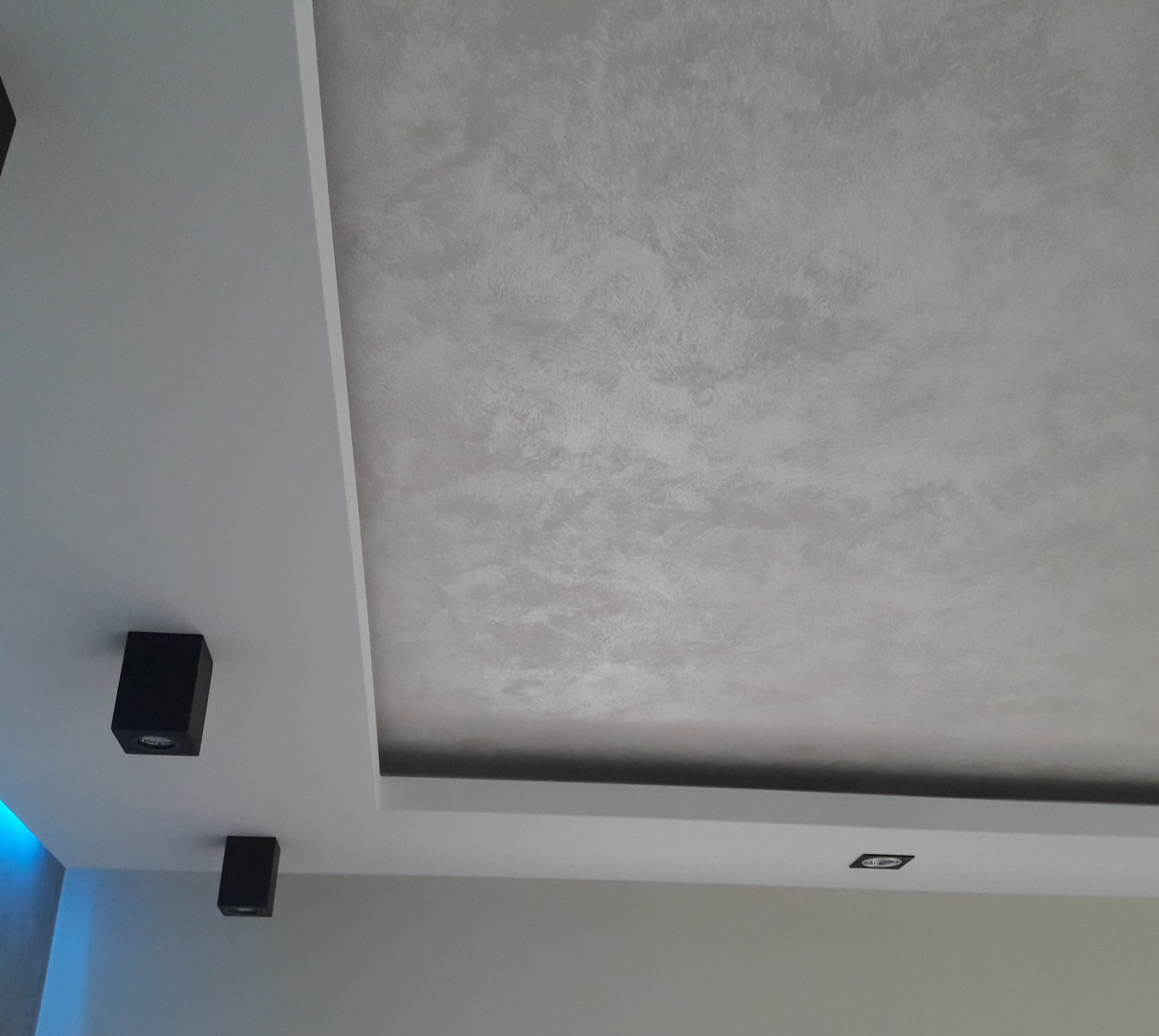 Fragment sufitu z dekoracyjną strukturą w odcieniach szarości, obniżony z wbudowanym oświetleniem punktowym i dwoma czarnymi reflektorami.