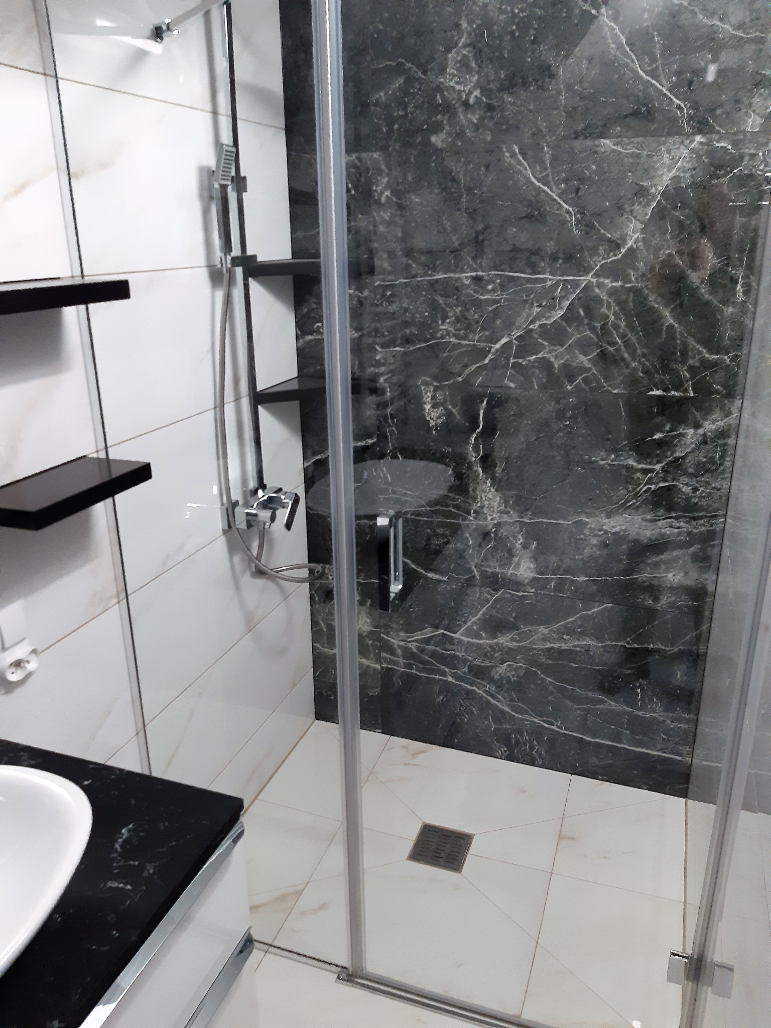 Nowoczesna łazienka z kabiną prysznicową typu walk-in, wykończona białymi i czarnymi płytkami imitującymi marmur, z czarnymi półkami i armaturą w kolorze chromu.