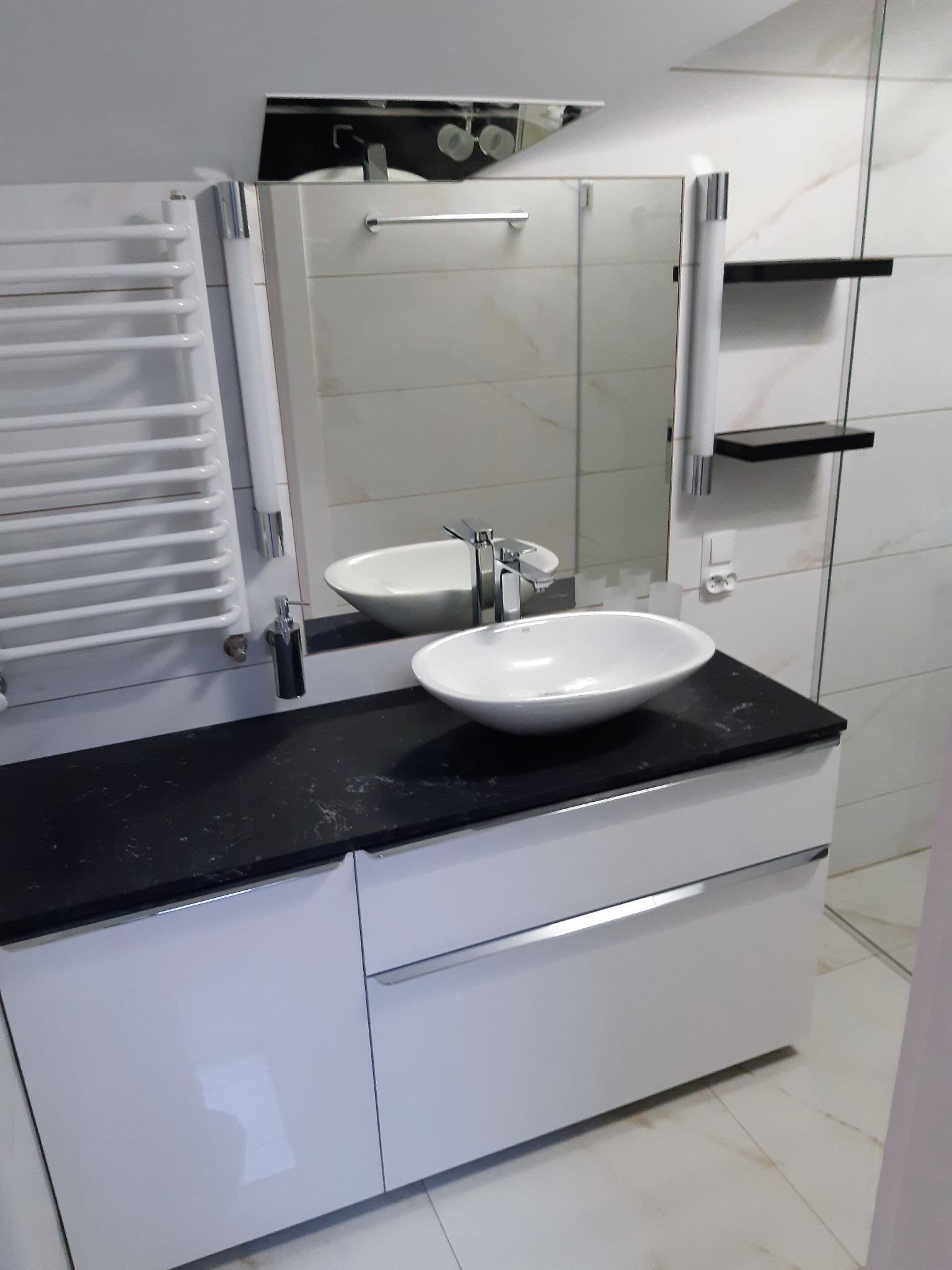 Nowoczesna łazienka z owalną umywalką nablatową, czarnym blatem i białymi szafkami pod umywalką, widoczny grzejnik drabinkowy i lustro z oświetleniem.