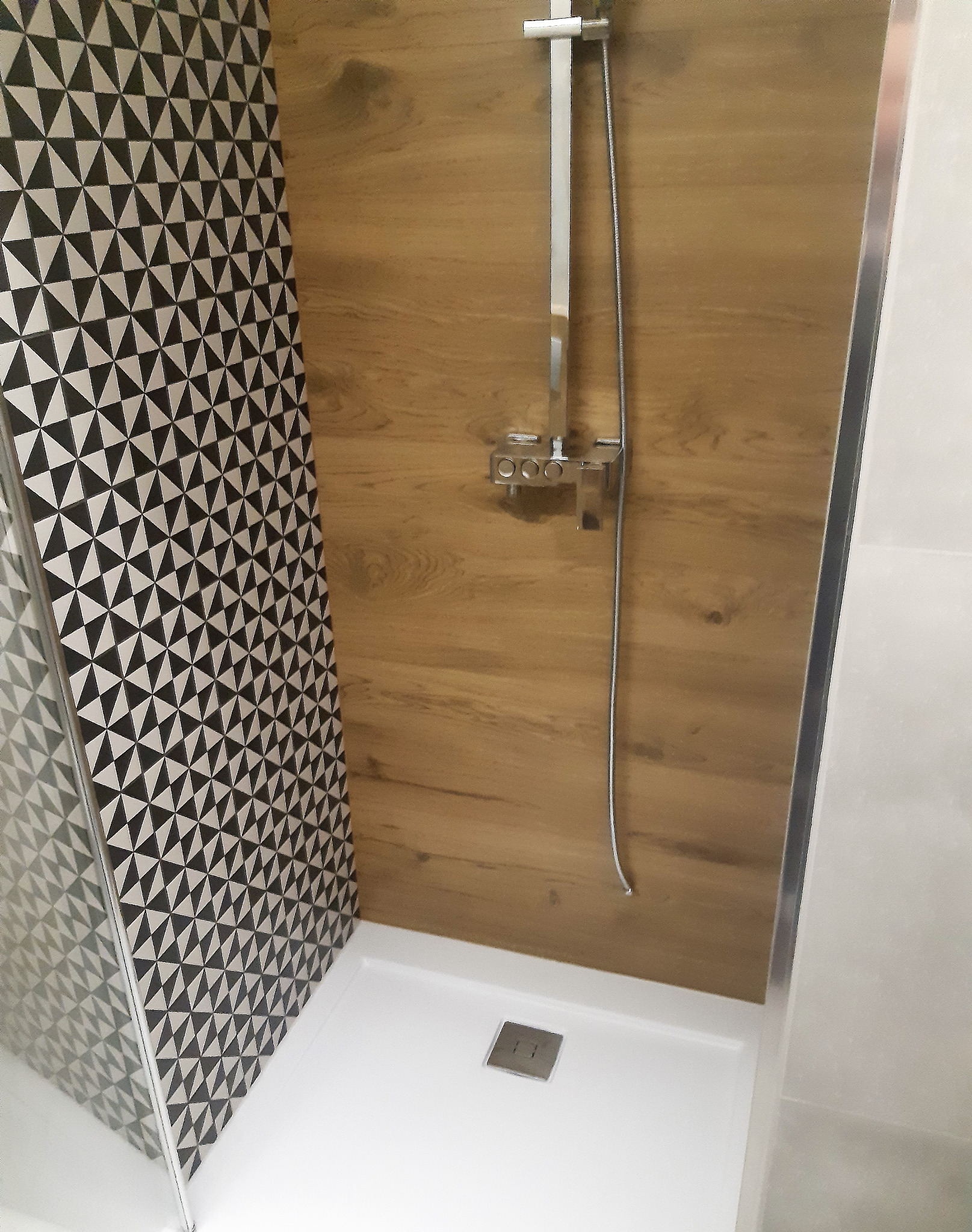 Kabina prysznicowa z panelami imitującymi drewno i geometryczną tapetą, chromowana armatura, biały brodzik z odpływem.