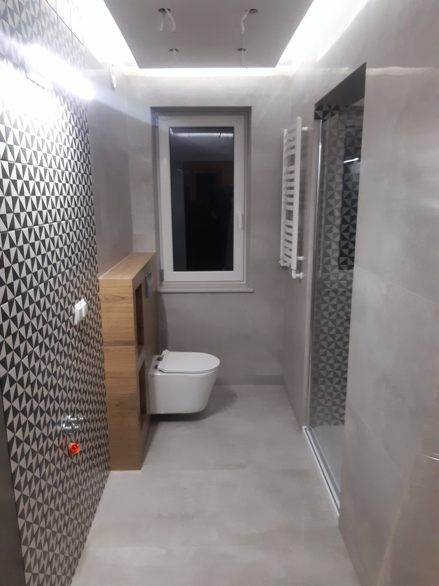 Wykończona łazienka z geometryczną mozaiką na ścianie, podwieszaną toaletą, grzejnikiem drabinkowym i kabiną prysznicową ze szklanymi drzwiami, oświetlenie sufitowe LED.