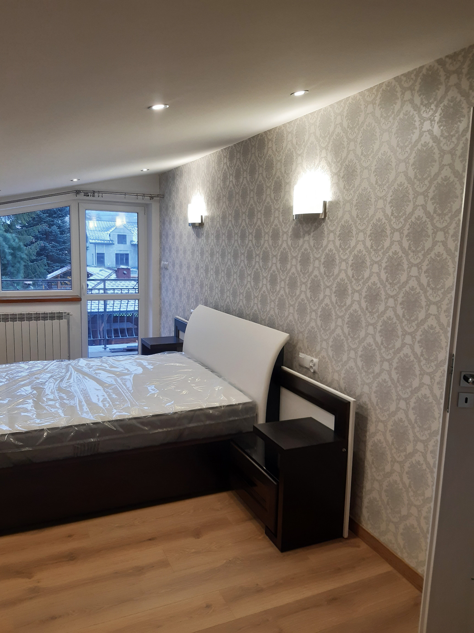 Pokój z łóżkiem w folii ochronnej, ściana w jasne, wzorzyste tapety, drewniana podłoga, lampki ścienne i okno z widokiem na zimowy krajobraz.