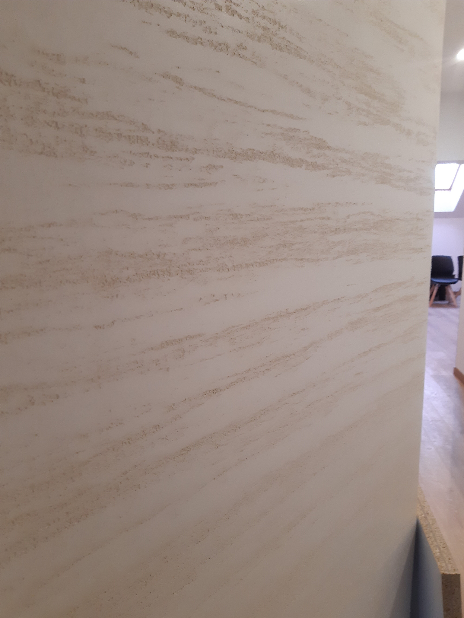 Ściana z beżową, dekoracyjną strukturą przypominającą piaskowiec, widoczna z bliska, z fragmentem wnętrza w tle.