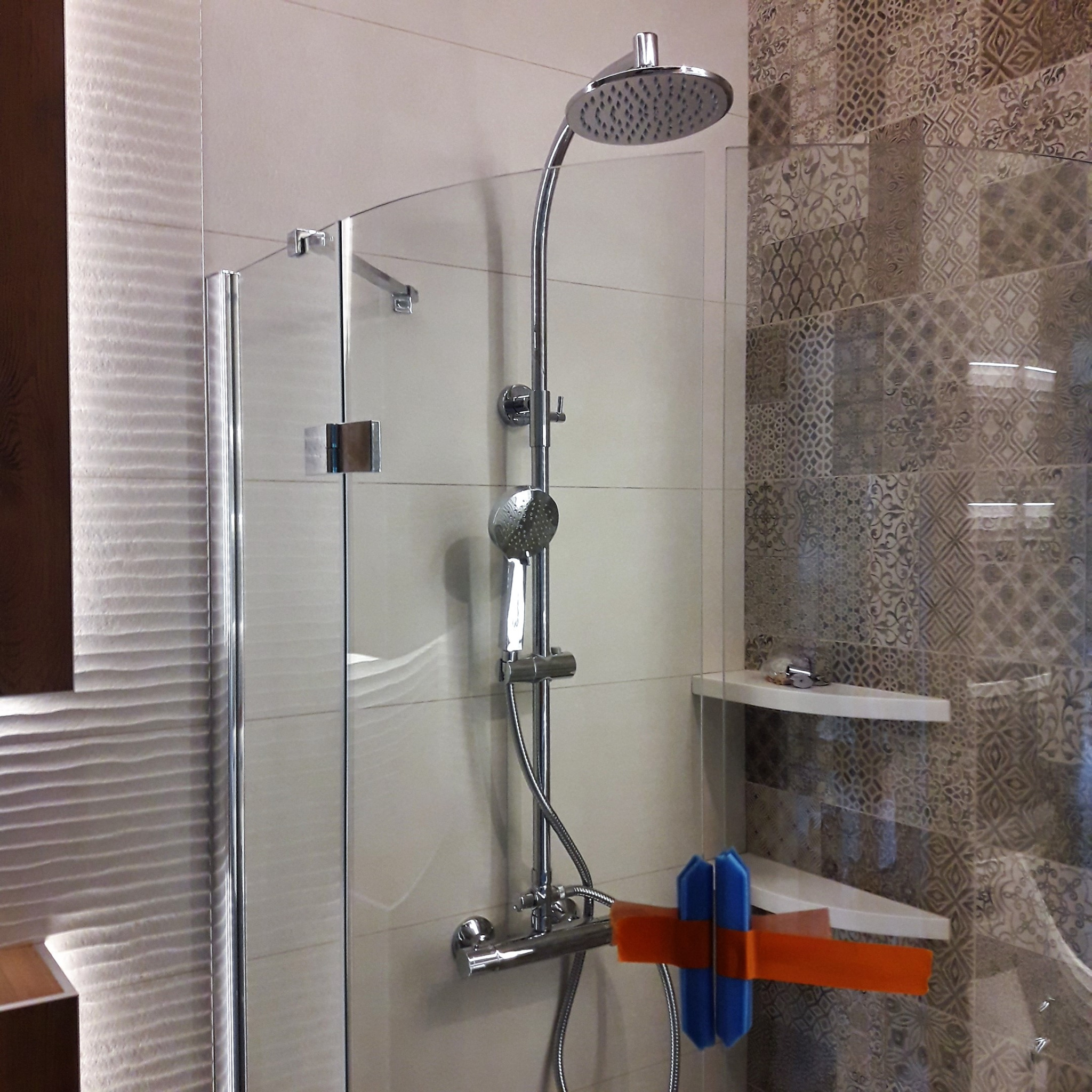 Nowoczesna kabina prysznicowa z deszczownicą i półkami, wykończona płytkami w różnych wzorach i fakturach. Na baterii prysznicowej zawieszona ściągaczka do wody z niebieskimi końcówkami...