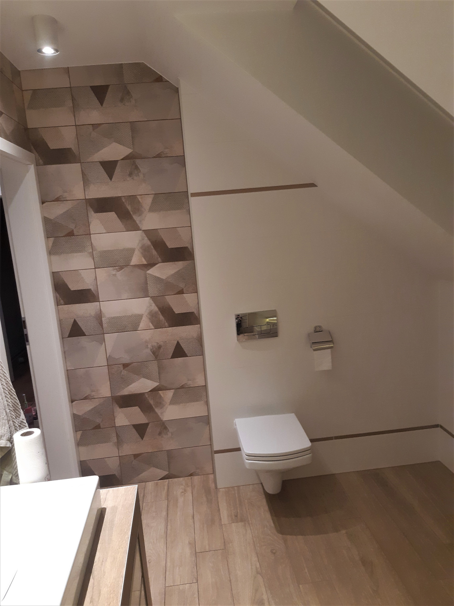 Ukończona łazienka na poddaszu z toaletą podwieszaną, drewnopodobną podłogą i geometrycznymi płytkami na ścianie przy wejściu.