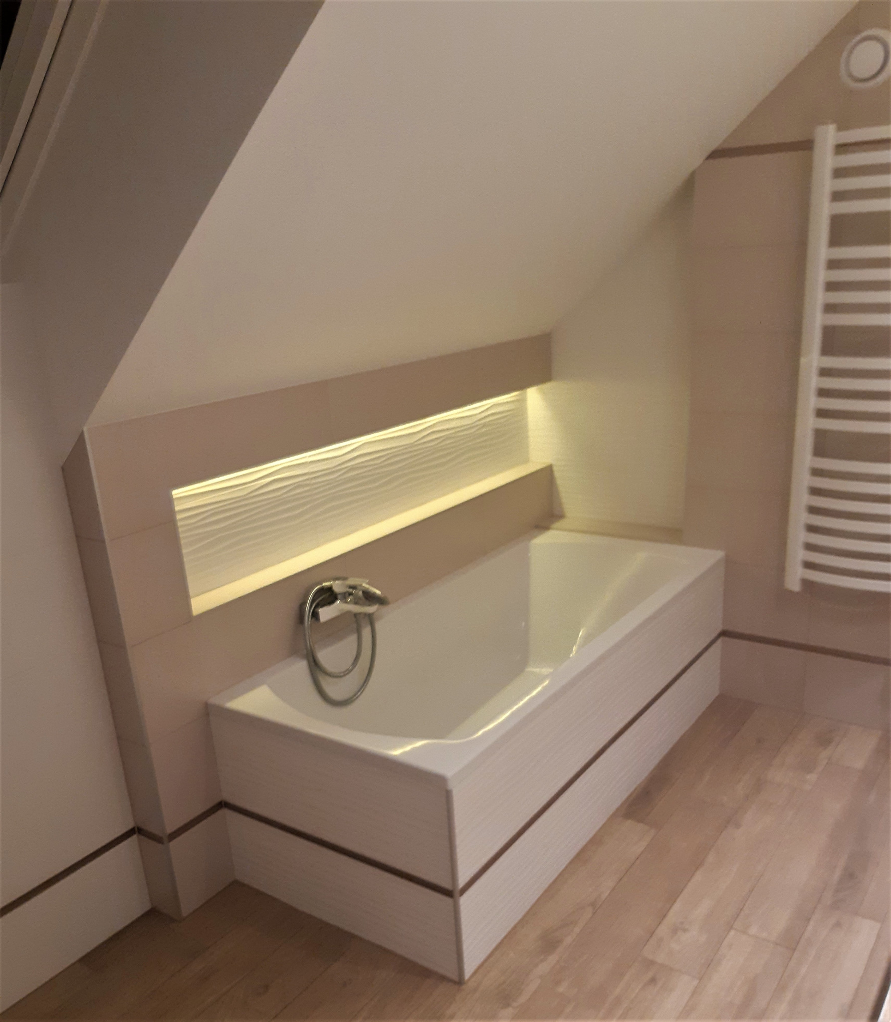 Wanna w obudowie z płytek, podświetlona dekoracyjna ściana w niszy, grzejnik drabinkowy, łazienka na poddaszu ze skośnym sufitem, drewnopodobna podłoga.