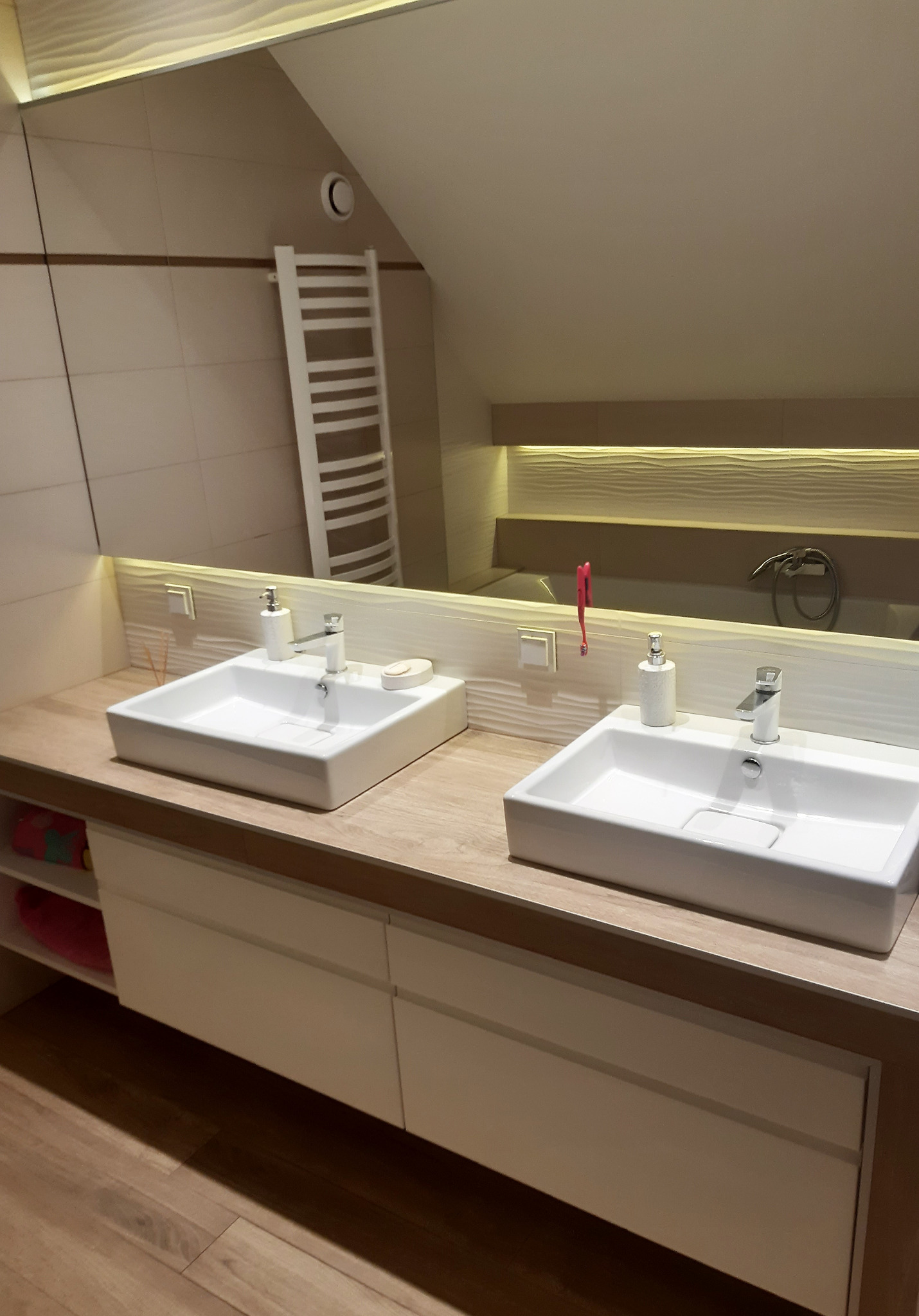 Umywalki nablatowe w nowoczesnej łazience na poddaszu z drewnianym blatem i białymi szafkami; widoczne akcesoria łazienkowe i grzejnik drabinkowy w odbiciu lustra.