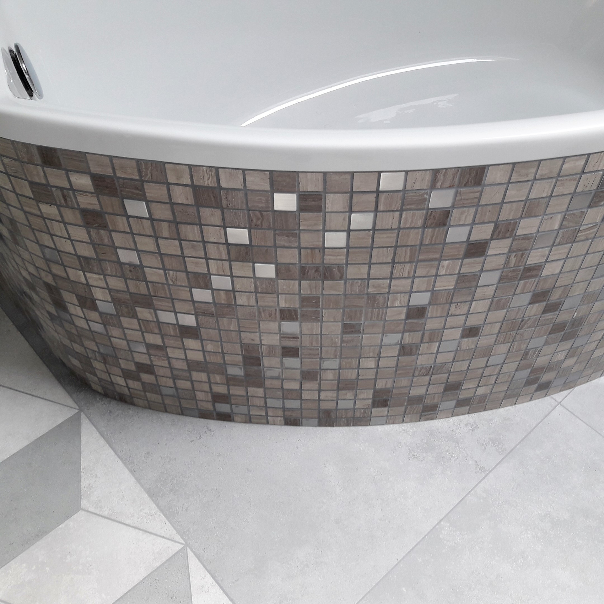 Obudowa wanny wykończona mozaiką w odcieniach brązu i szarości, uzupełniona jasną, geometryczną posadzką z płytek.