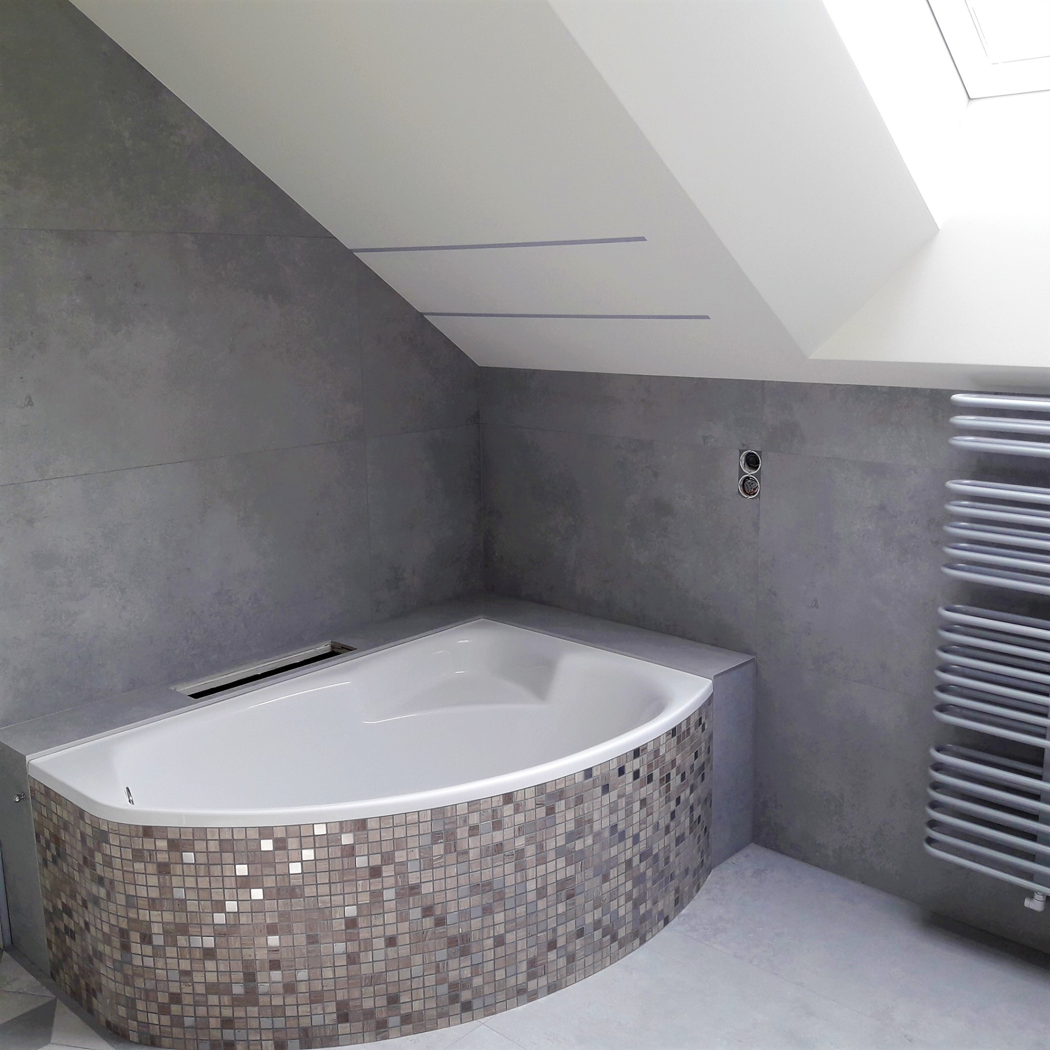 Narożna wanna obudowana mozaiką w szaro-beżowych odcieniach, szare płytki na ścianach w łazience na poddaszu ze skośnym sufitem i grzejnikiem drabinkowym.