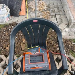 Electro-Stach Janusz Stachura - Miernik rezystancji uziemienia, ustawiony na plastikowym krześle na tle placu budowy z kostką brukową i ziemią. Widoczny przewód uziemiający z żółtą klamrą.