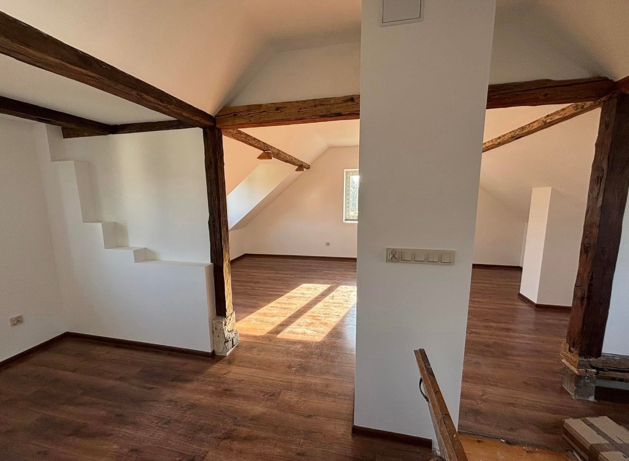 Wnętrze poddasza z drewnianymi belkami konstrukcyjnymi, widoczne gniazdka elektryczne na ścianie, drewniana podłoga i schody.
