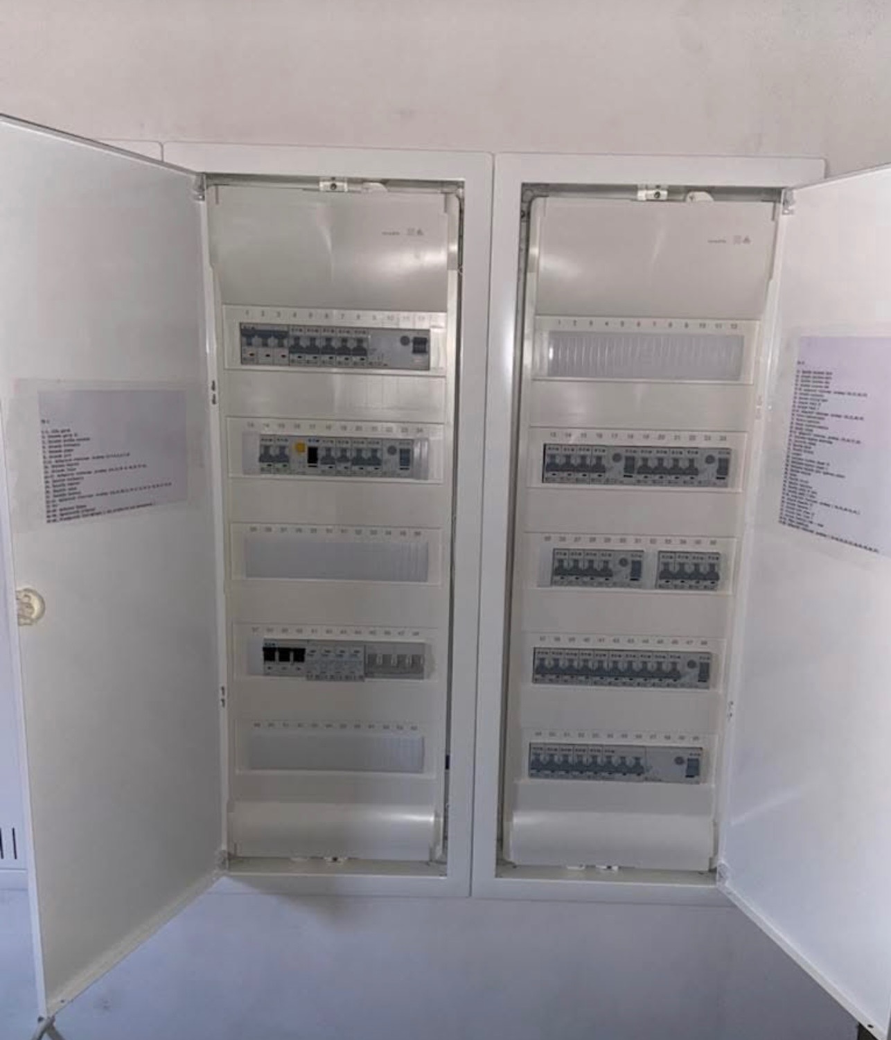 Otwarta szafa rozdzielcza z widocznymi modułowymi zabezpieczeniami, wyłącznikami i listwami zaciskowymi w białej obudowie, z dokumentacją techniczną po prawej stronie.