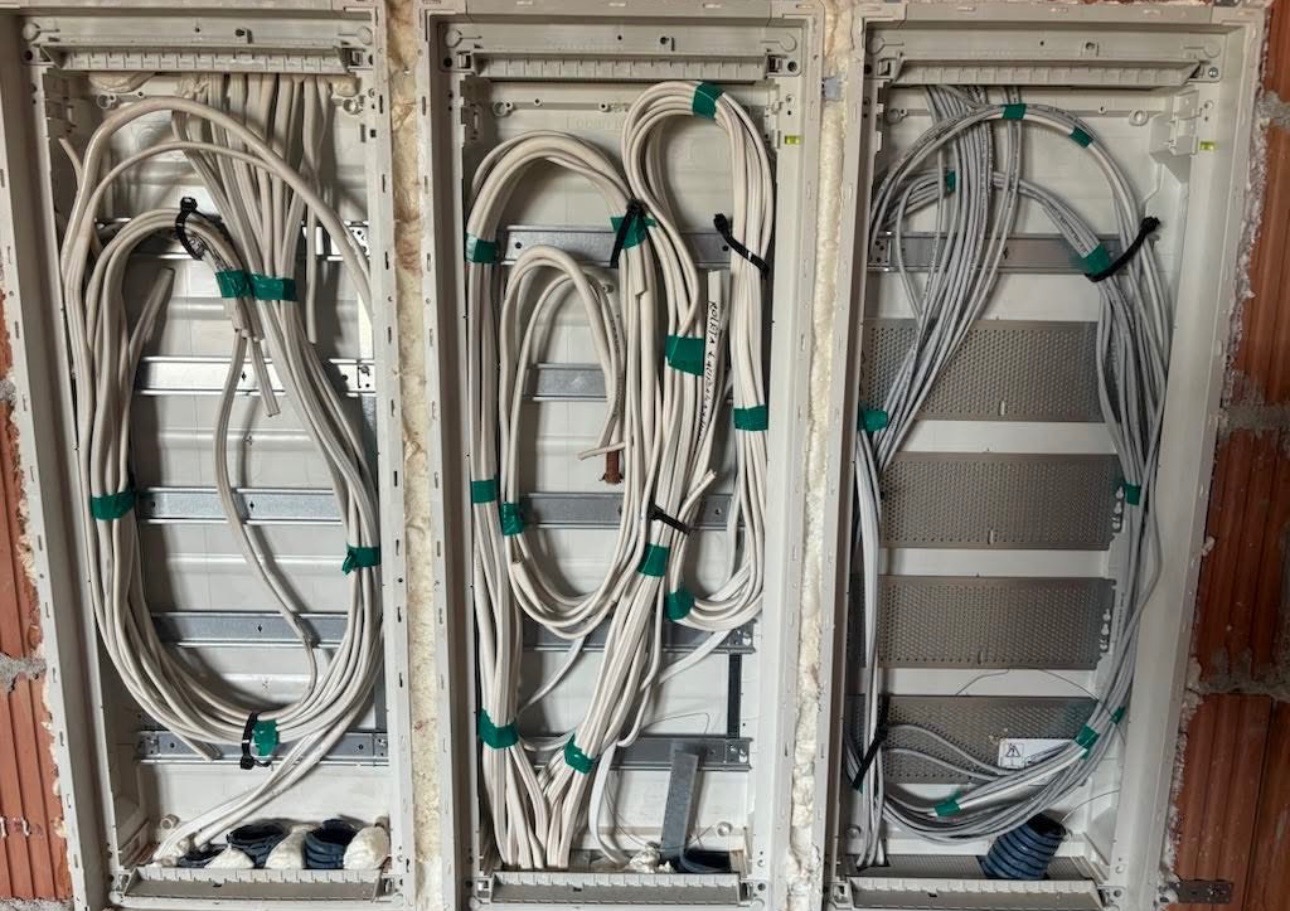 Trzy puste skrzynki rozdzielcze z poprowadzonymi kablami elektrycznymi, zabezpieczonymi zielonymi i czarnymi opaskami, zamontowane w ścianie z cegły.