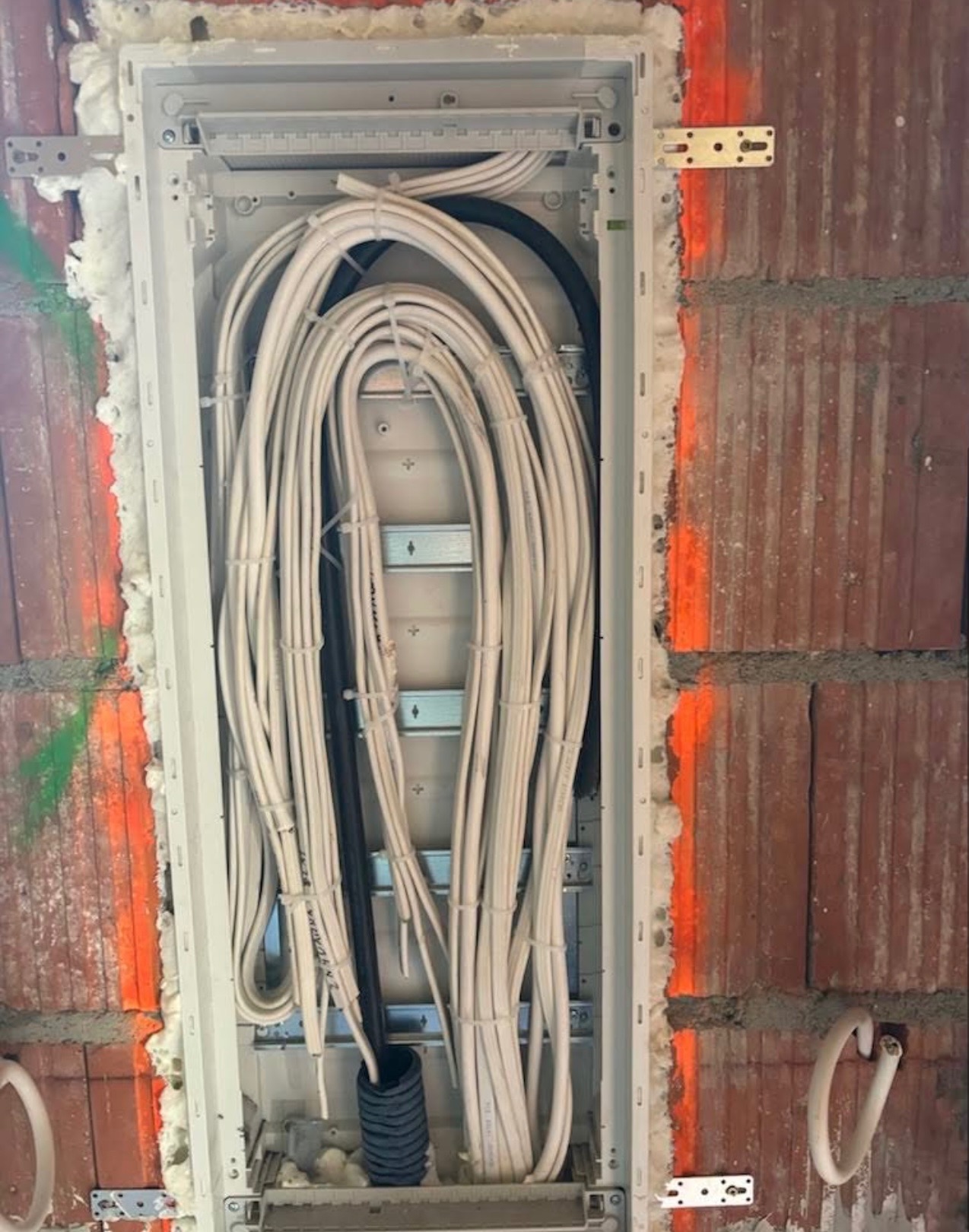 Rozdzielnica elektryczna w trakcie montażu w surowej ścianie z cegły, z widocznymi kablami i pianką montażową, zabezpieczona pomarańczową farbą.