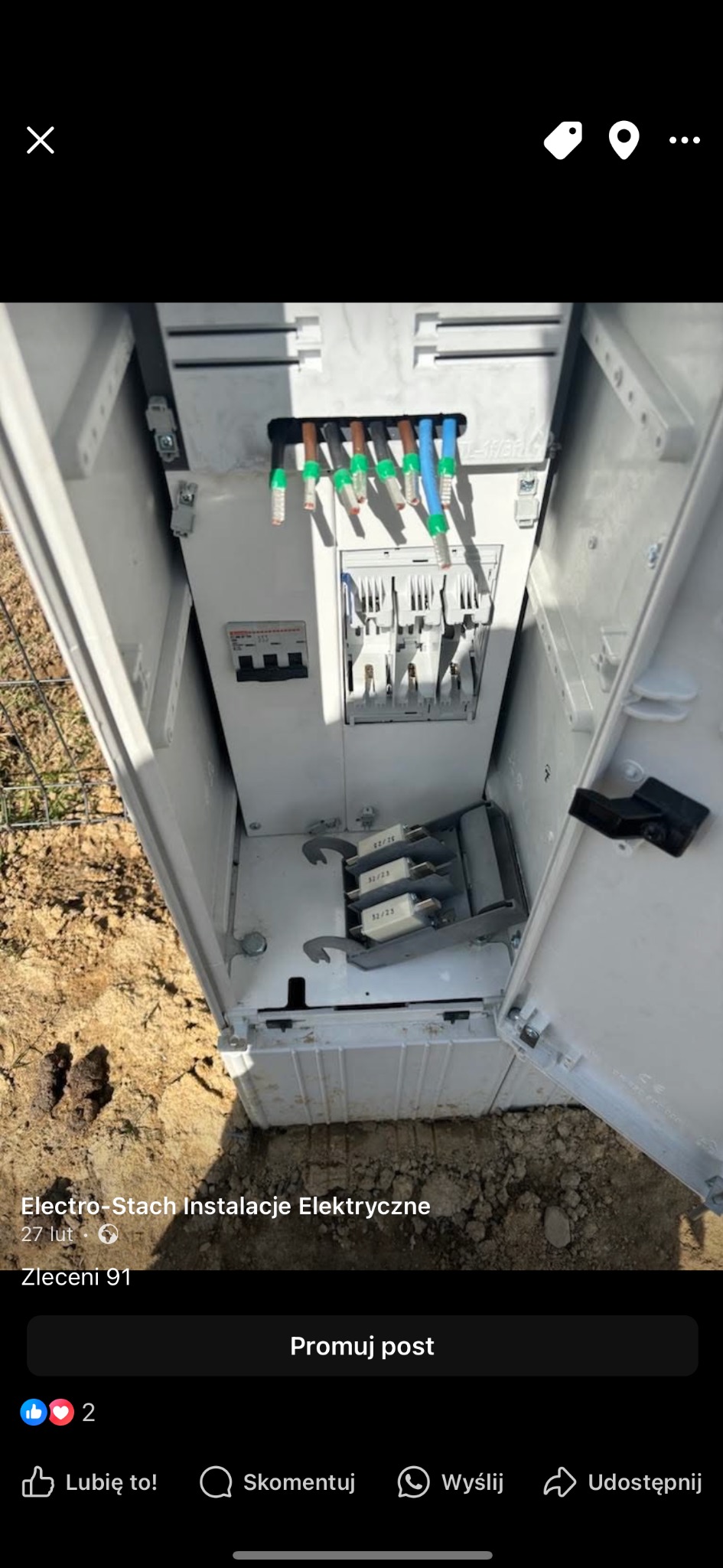 Otwarta skrzynka elektryczna z widocznymi przewodami i bezpiecznikami, zamontowana na zewnątrz, z ziemią w tle.