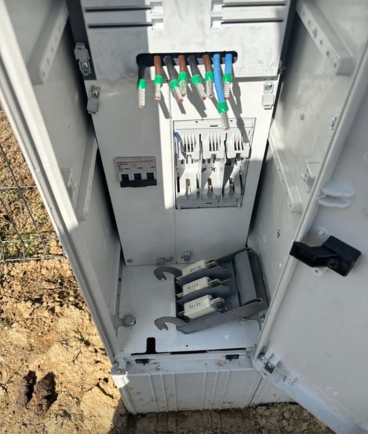 Otwarta skrzynka elektryczna z widocznymi przewodami, bezpiecznikami i elementami złącznymi, umieszczona na ziemi.