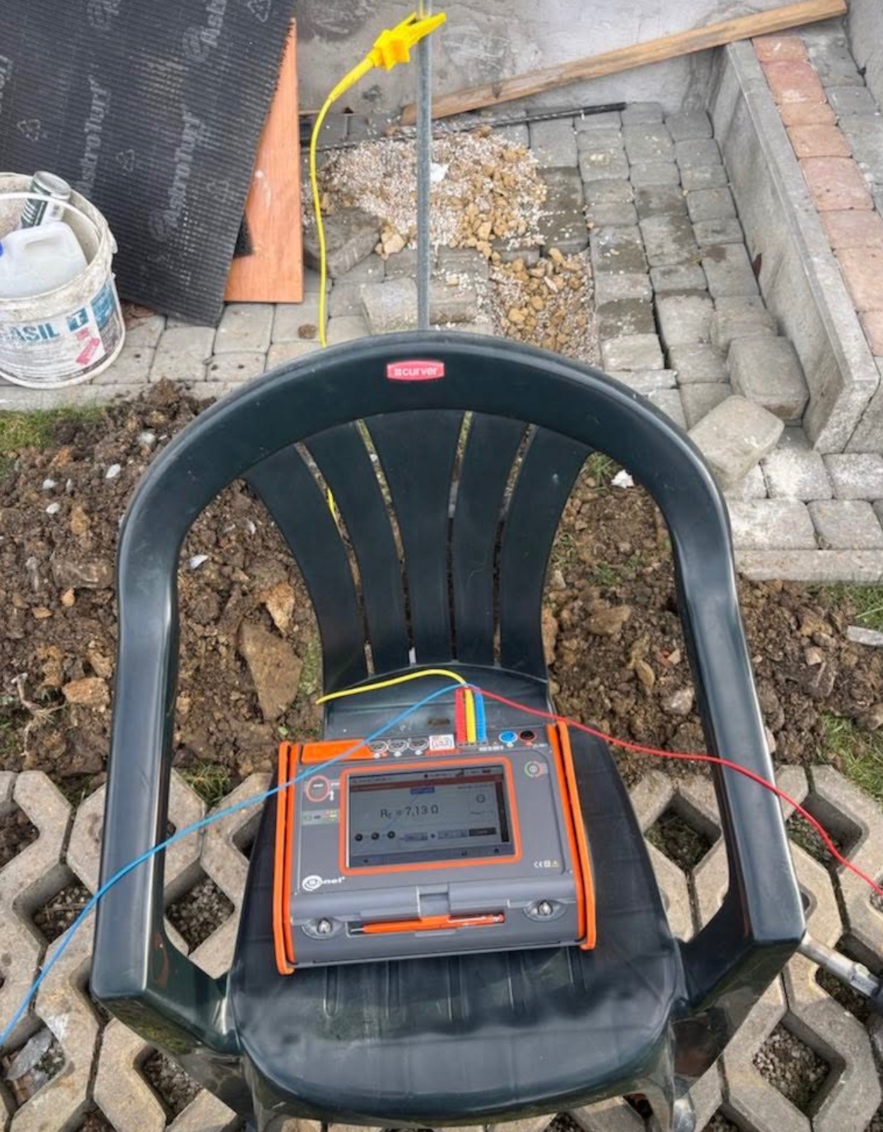 Miernik rezystancji uziemienia, ustawiony na plastikowym krześle na tle placu budowy z kostką brukową i ziemią. Widoczny przewód uziemiający z żółtą klamrą.