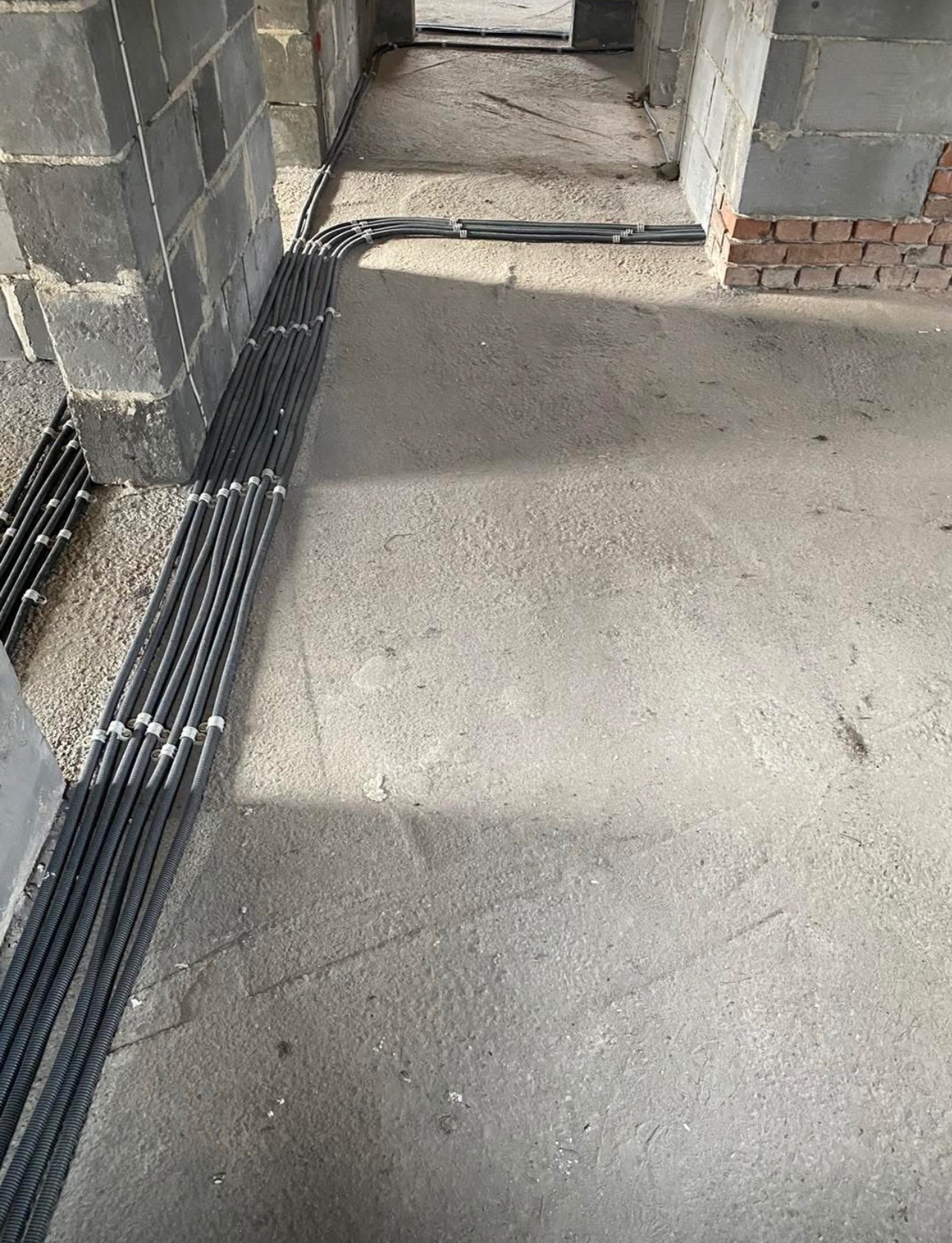 Wiązka czarnych, karbowanych rur instalacyjnych prowadzona wzdłuż surowej ściany z betonowych bloków, mocowana białymi uchwytami, na szarym, betonowym podłożu w stanie surowym.