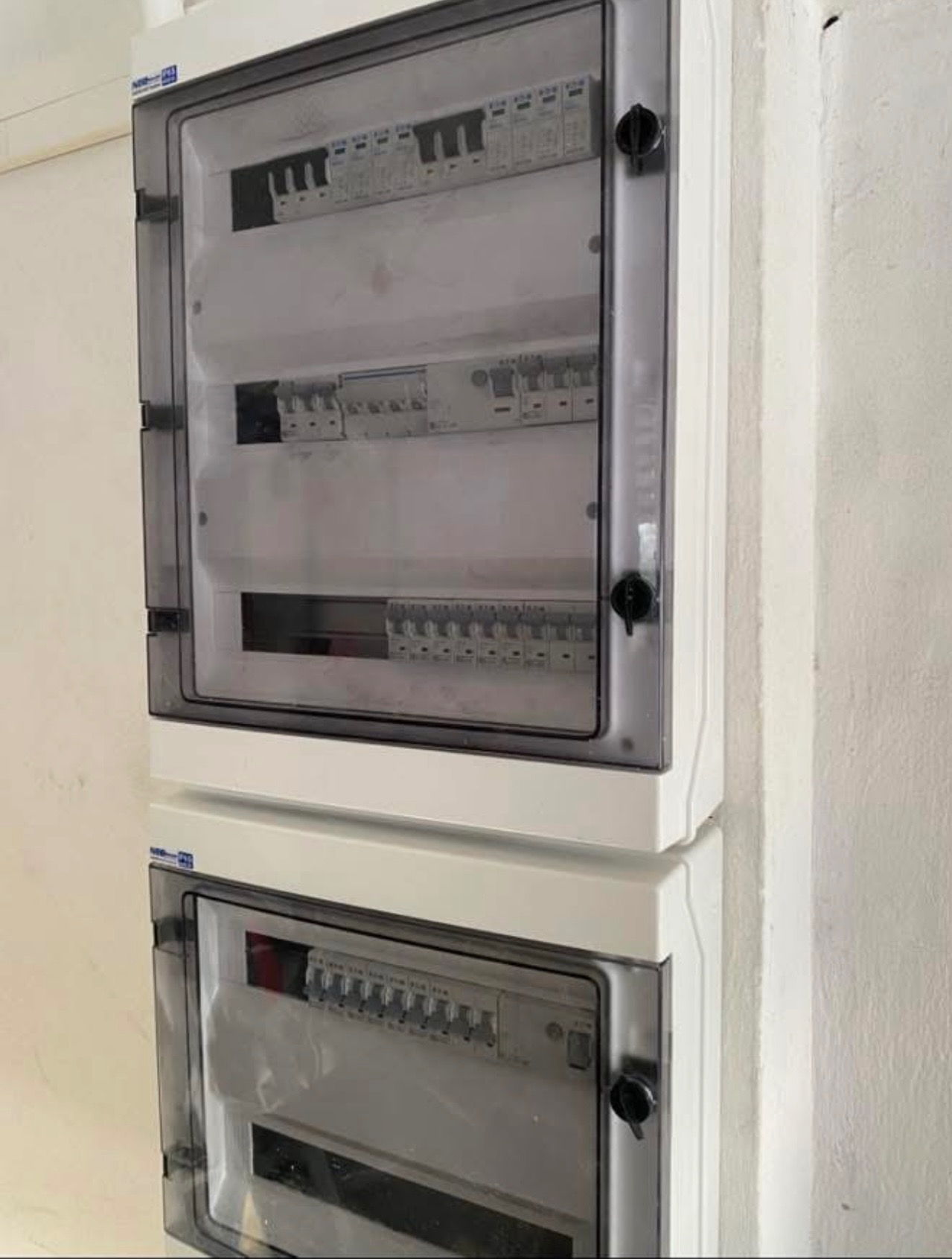 Dwa białe panele elektryczne z przezroczystymi drzwiczkami, zamontowane jeden nad drugim na ścianie w Jordanowie. Widoczne zabezpieczenia i wyłączniki wewnątrz.