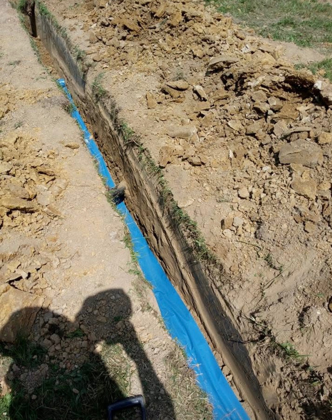 Wykopany wąski rów w ziemi, wyłożony niebieską folią ochronną, z widocznym cieniem osoby fotografującej i fragmentem szpadla.