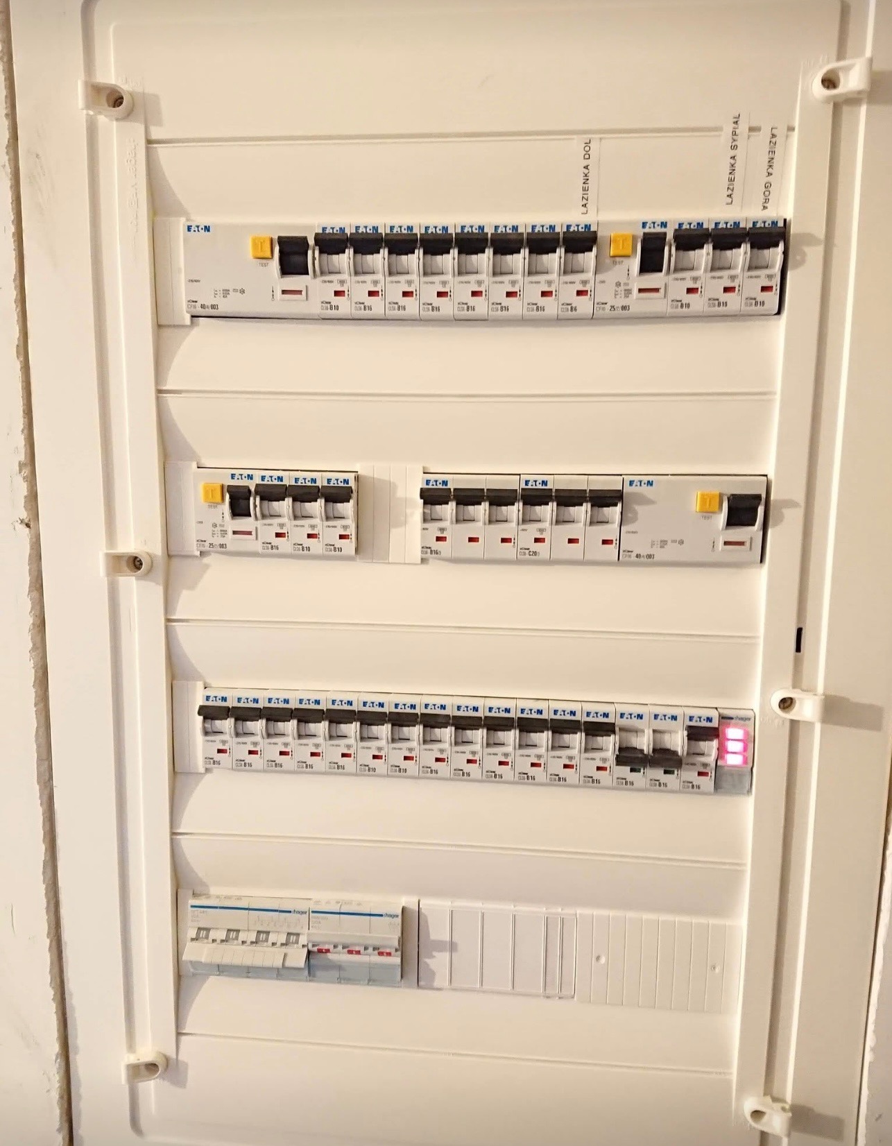 Rozdzielnica elektryczna z bezpiecznikami marki Eaton, opisana etykietami 'Łazienka Dol', 'Łazienka Sypial' i 'Łazienka Góra', jeden z bezpieczników włączony sygnalizuje działanie obwodu.