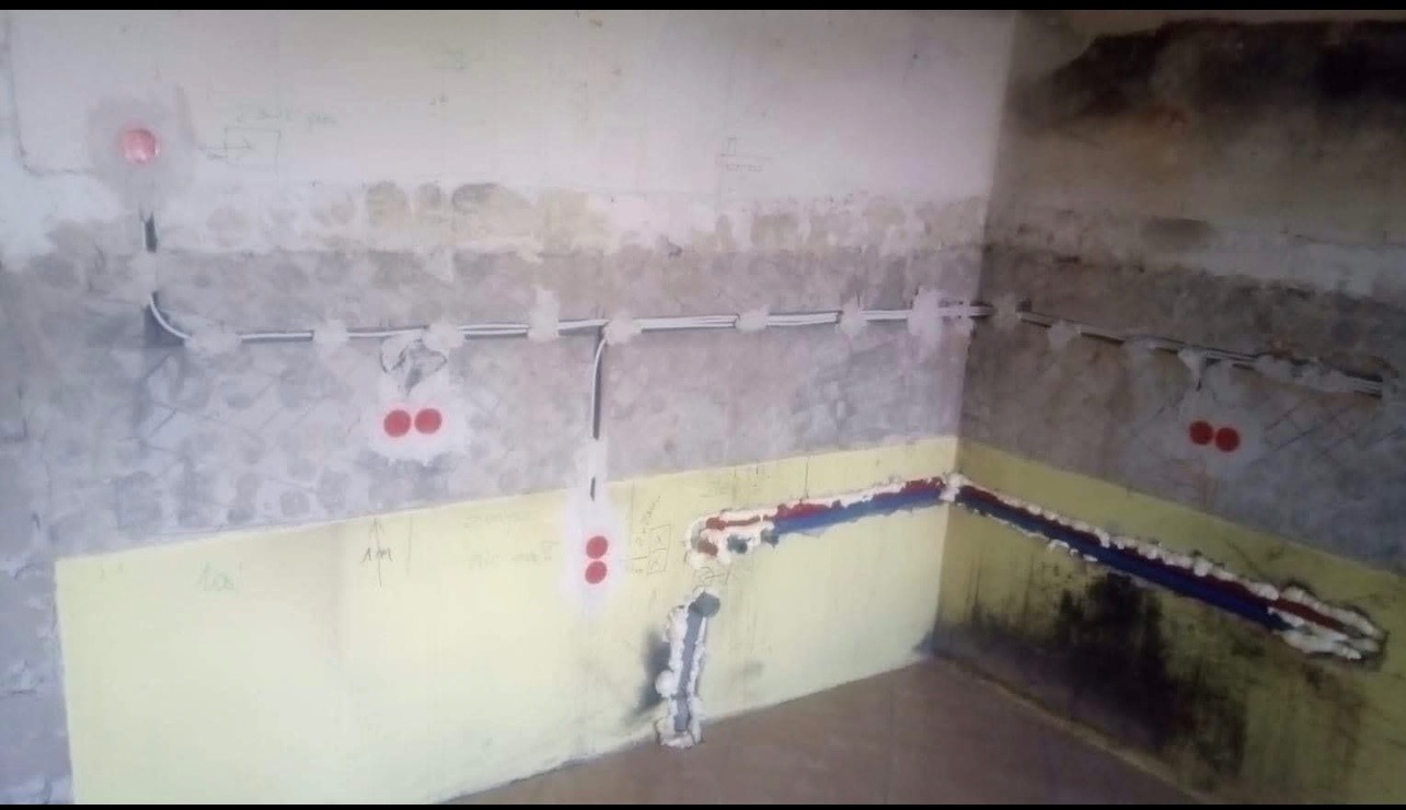 Wnętrze w trakcie remontu, widoczne nowe instalacje elektryczne i hydrauliczne w ścianach, zabezpieczone pianką montażową i puszkami elektrycznymi, surowe ściany z widocznymi śladami po pracach.