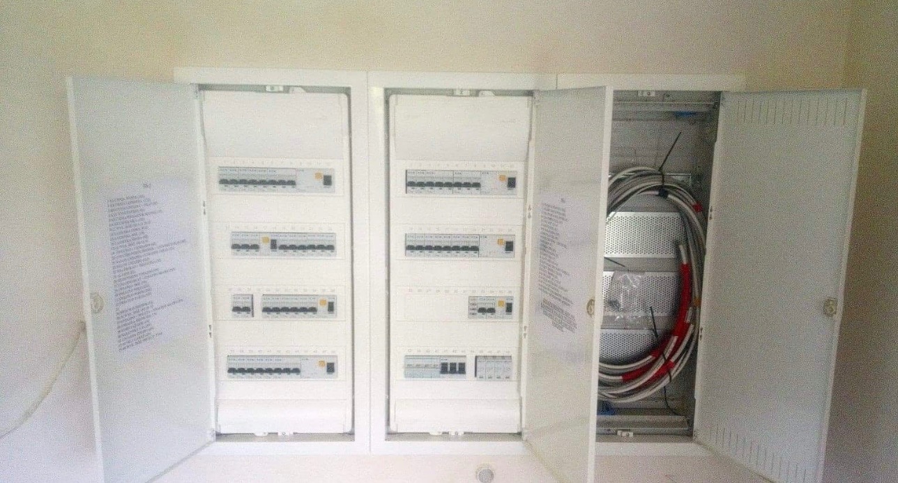 Otwarta skrzynka rozdzielcza z widocznymi bezpiecznikami i przewodami wewnątrz, z dokumentacją techniczną na wewnętrznej stronie drzwi.
