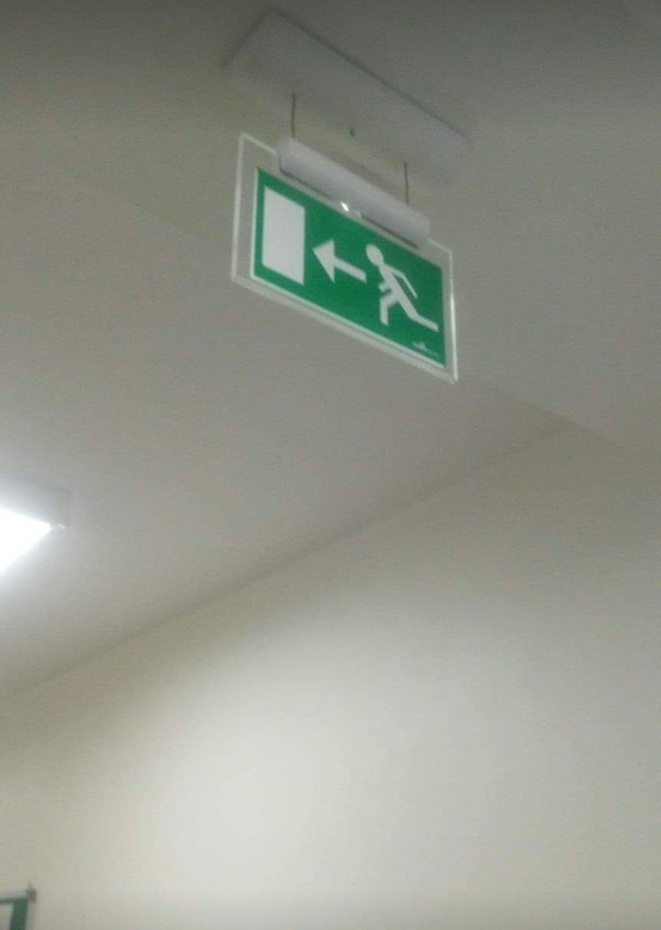 Podświetlany, zielony znak ewakuacyjny z piktogramem biegnącej osoby i strzałką w lewo, zamontowany na suficie.