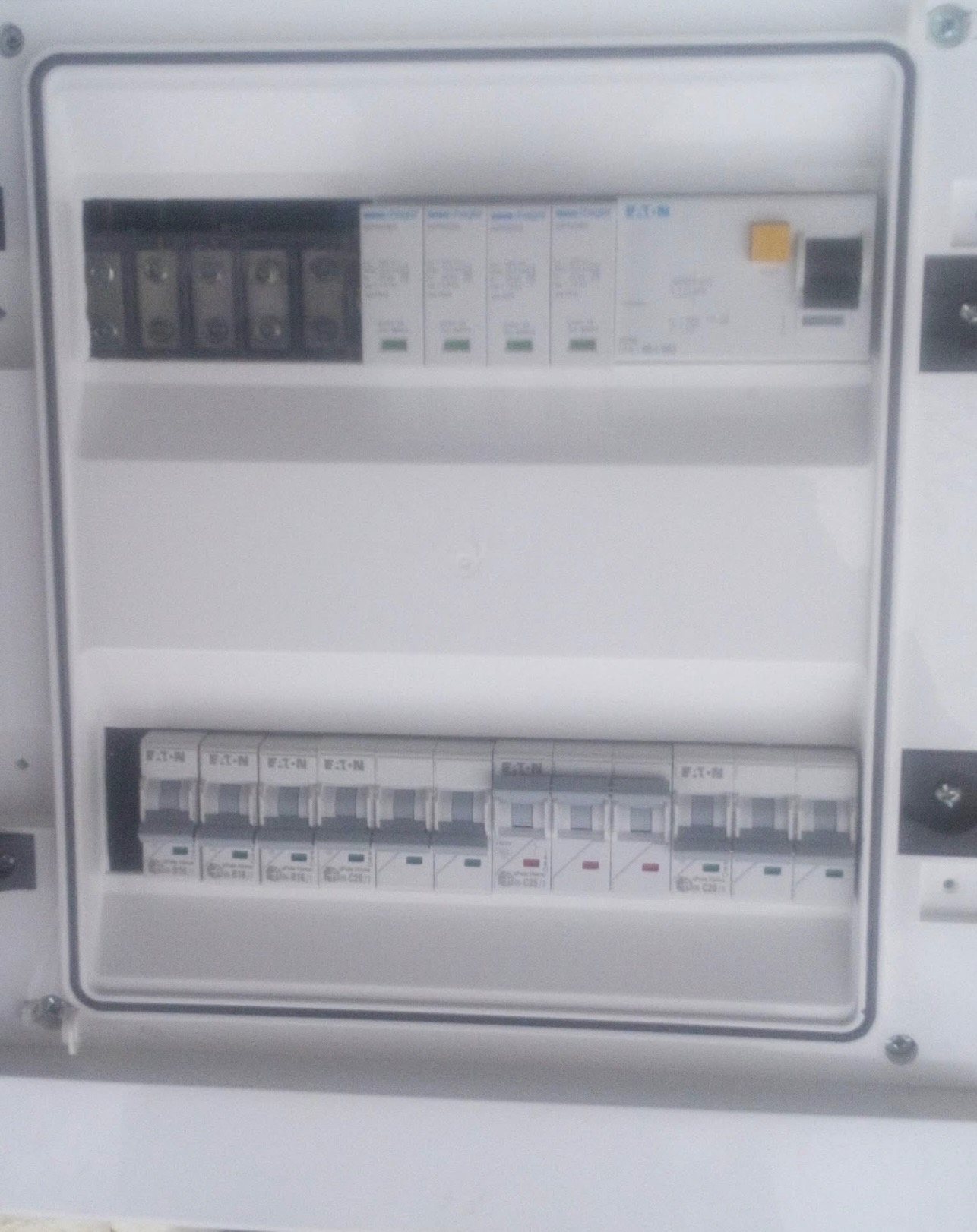 Otwarta, biała skrzynka elektryczna z widocznymi bezpiecznikami i wyłącznikami firmy Eaton. Górny rząd zawiera mniejsze wyłączniki, a dolny rząd większe, z czerwonymi i zielonymi wskaźnikami.