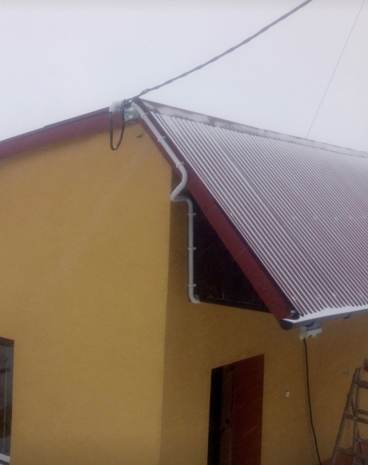 Żółty dom z bordowym dachem pokrytym śniegiem, widoczne rynny, przewody elektryczne i drabina oparta o ścianę w pochmurny dzień.