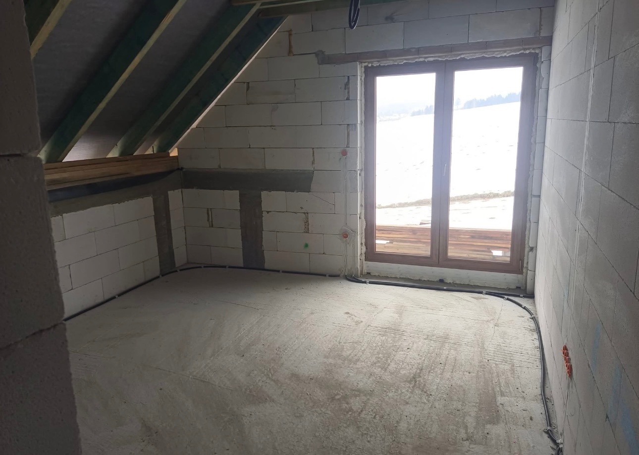 Surowy stan poddasza z widoczną instalacją elektryczną w peszlu przy podłodze i puszkami w ścianach z betonu komórkowego, okno dachowe z widokiem na zaśnieżone wzgórza.