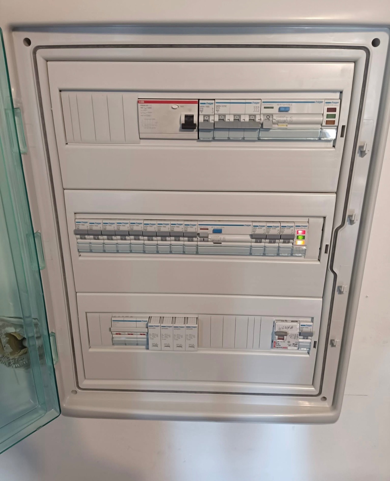 Otwarta skrzynka bezpiecznikowa z widocznymi wyłącznikami różnicowoprądowymi i nadprądowymi różnych typów i producentów, zamontowana na białej ścianie.