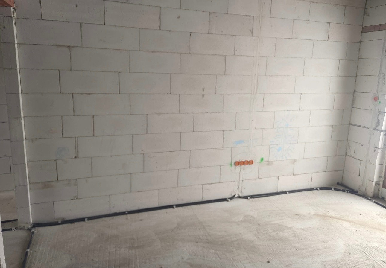 Surowa ściana z betonu komórkowego z widocznymi, prowadzonymi po podłodze i przymocowanymi do ściany przewodami elektrycznymi oraz puszkami instalacyjnymi w kolorze pomarańczowym.