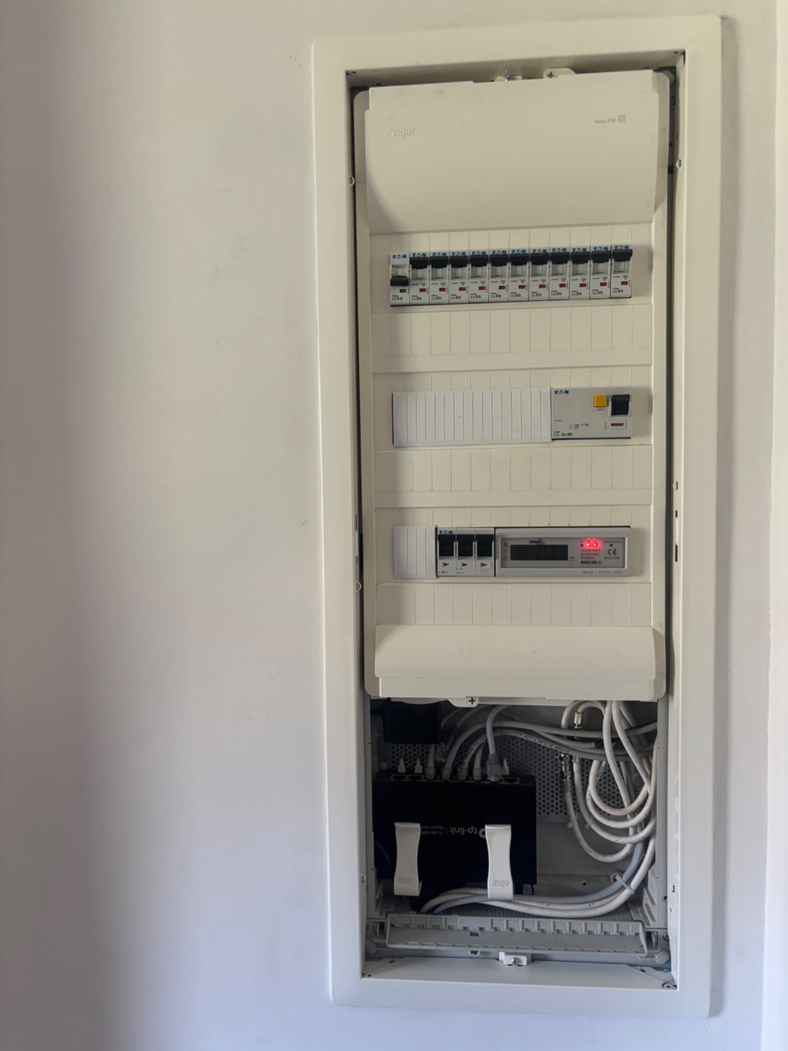 Otwarta skrzynka rozdzielcza wnękowa z widocznymi bezpiecznikami, licznikiem energii elektrycznej i modułem TP-Link, okablowanie w kolorze białym, zamontowana na białej ścianie.